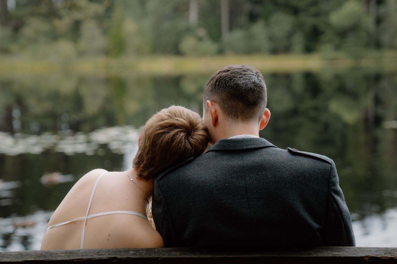 A couple in wedding attire sits on a bench facing a serene lake, the bride resting her head on the groom's shoulder.
