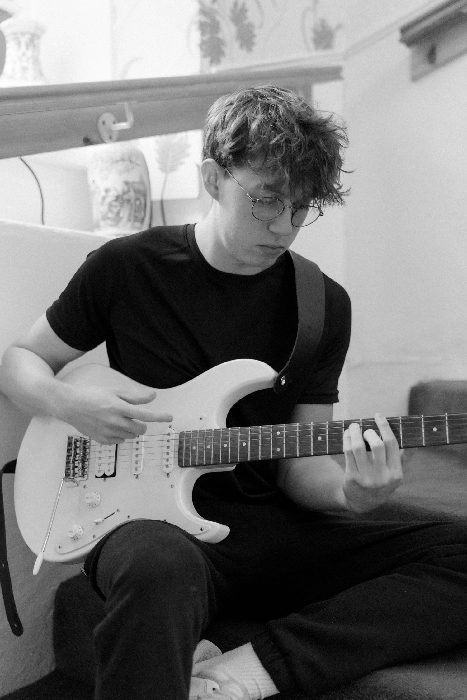 Young man with glasses playing electric guitar while seated, focused on his playing, black and white photo.