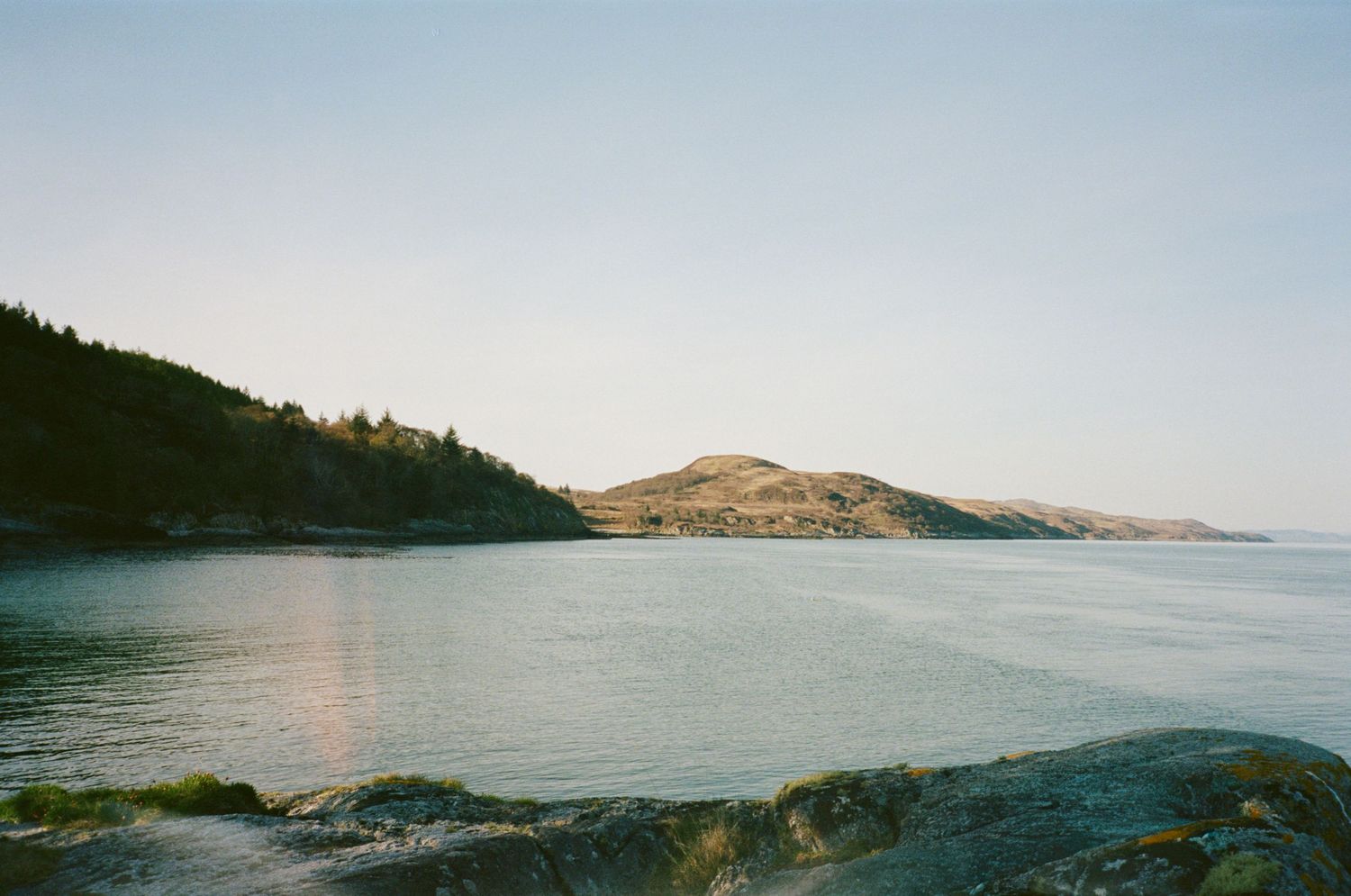 Scenic coastal landscape with calm blue waters, rocky shoreline, evergreen trees, and a rolling hill under clear sky.