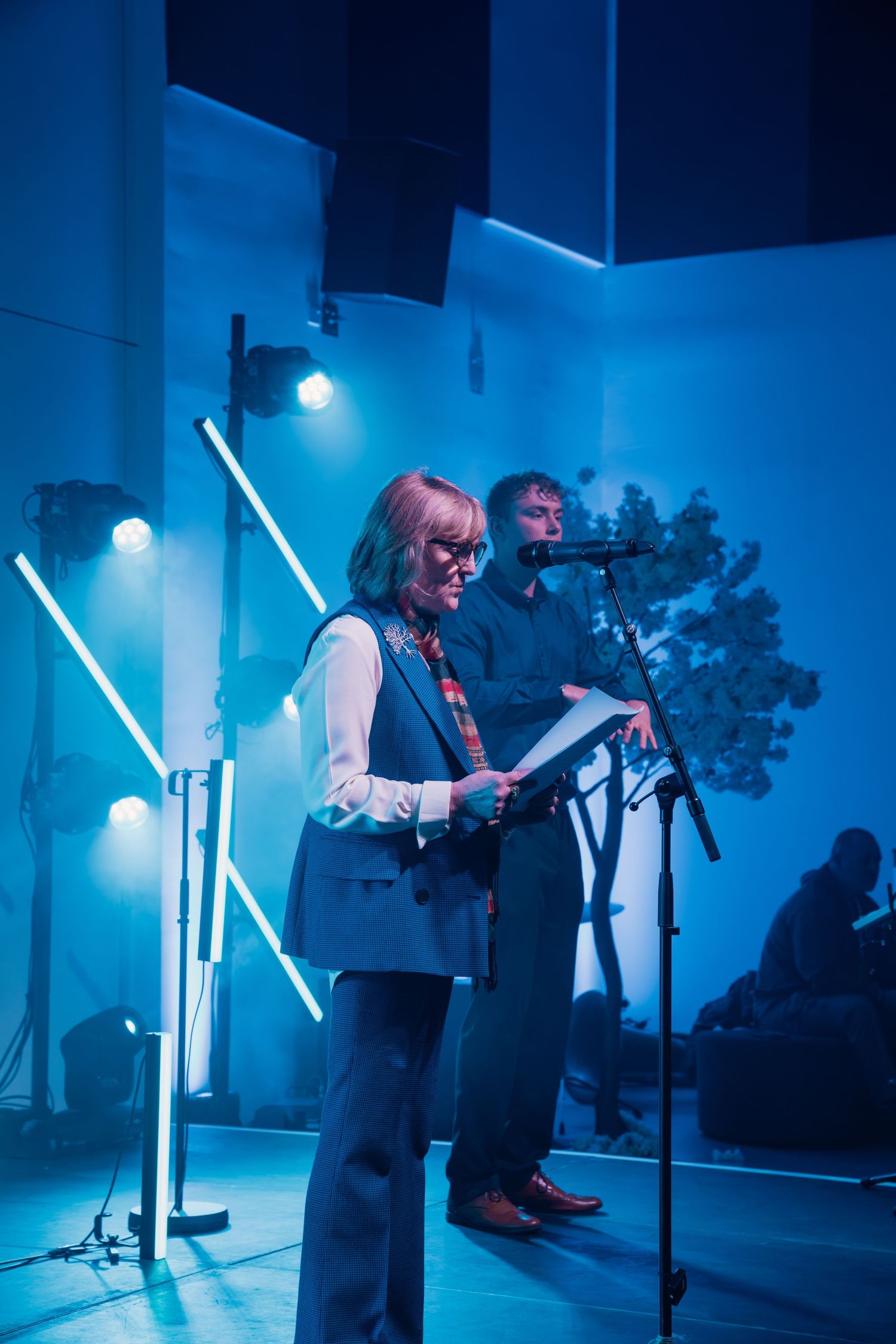 Two speakers present on a blue-lit stage with microphone, neon lights, and decorative tree backdrop.