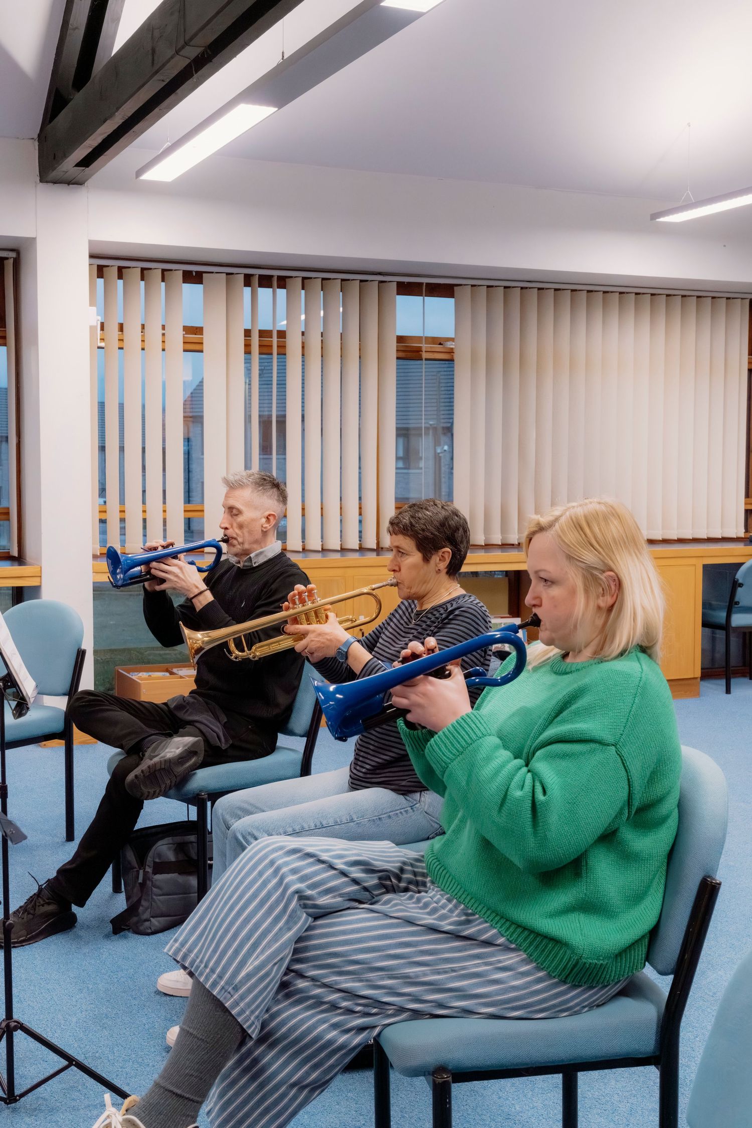 Three adults sit in chairs playing trumpets, including blue pластик models, during an indoor music rehearsal session.