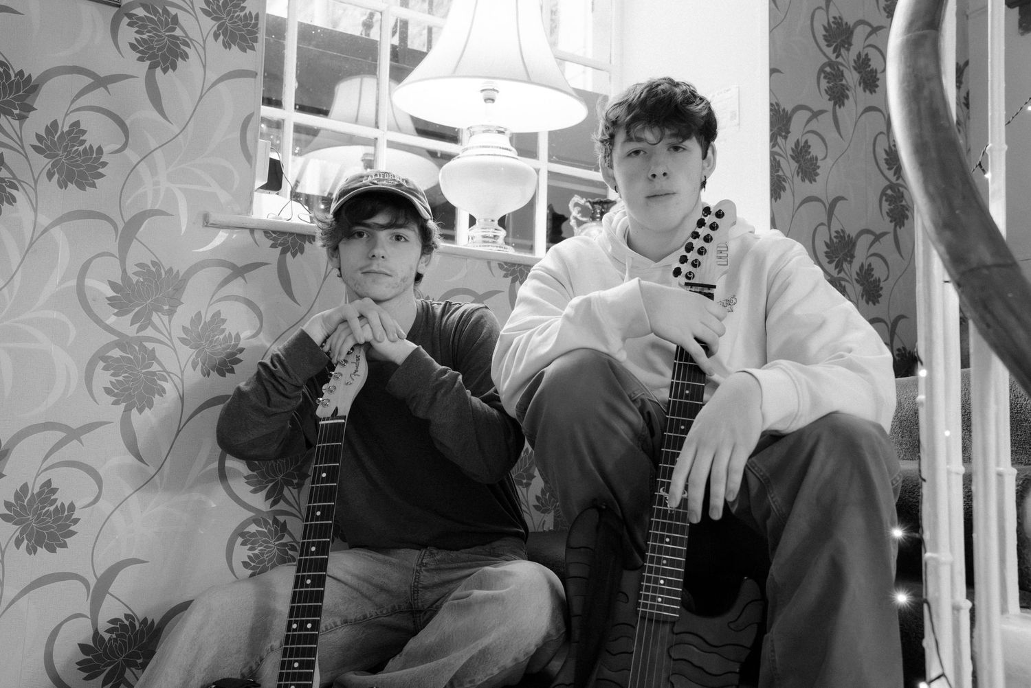 Two young men pose together in a black and white photo, one holding a guitar, against a floral wallpaper background.