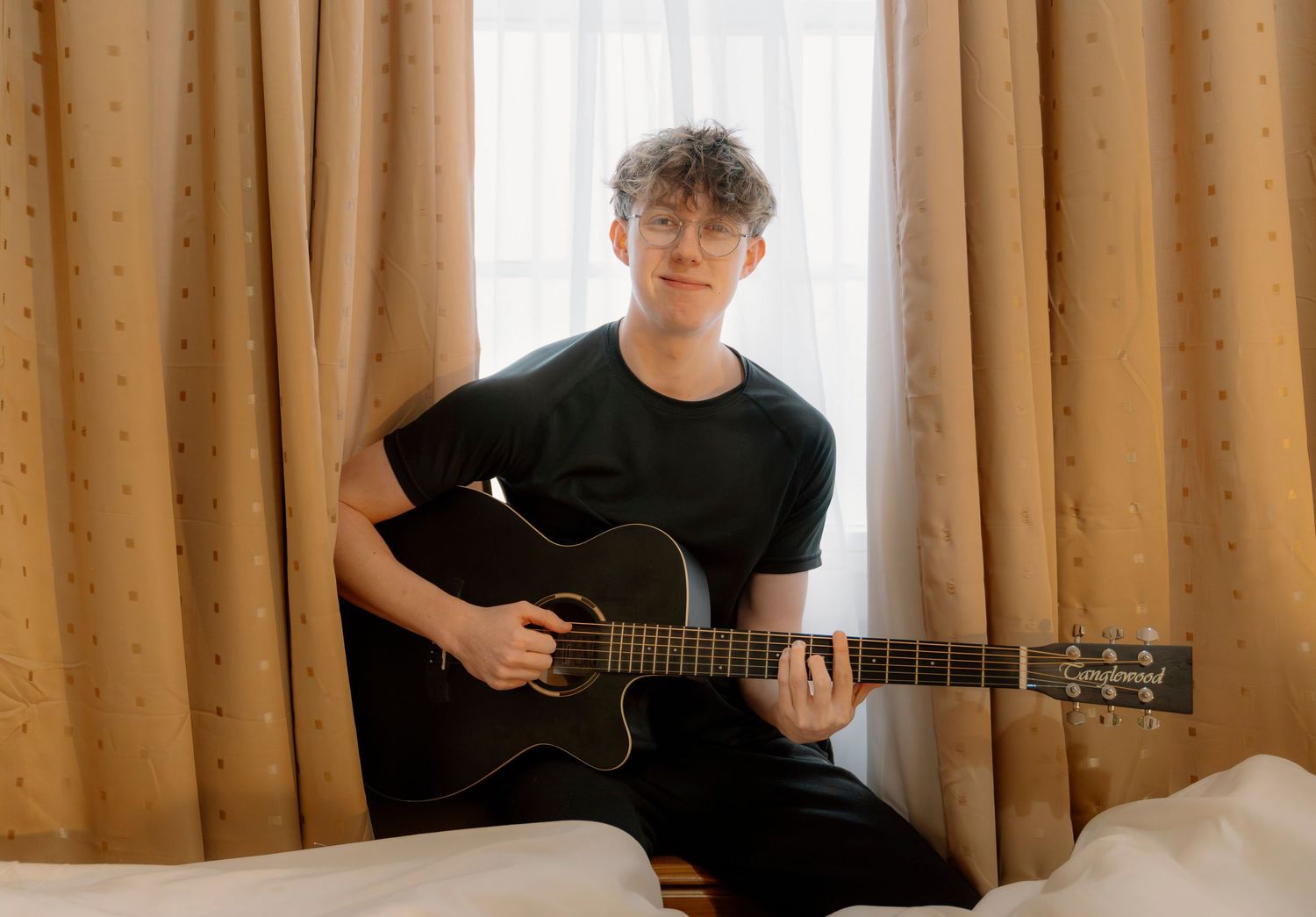 Young man with curly hair and glasses playing acoustic guitar while sitting on a bed near curtained windows.