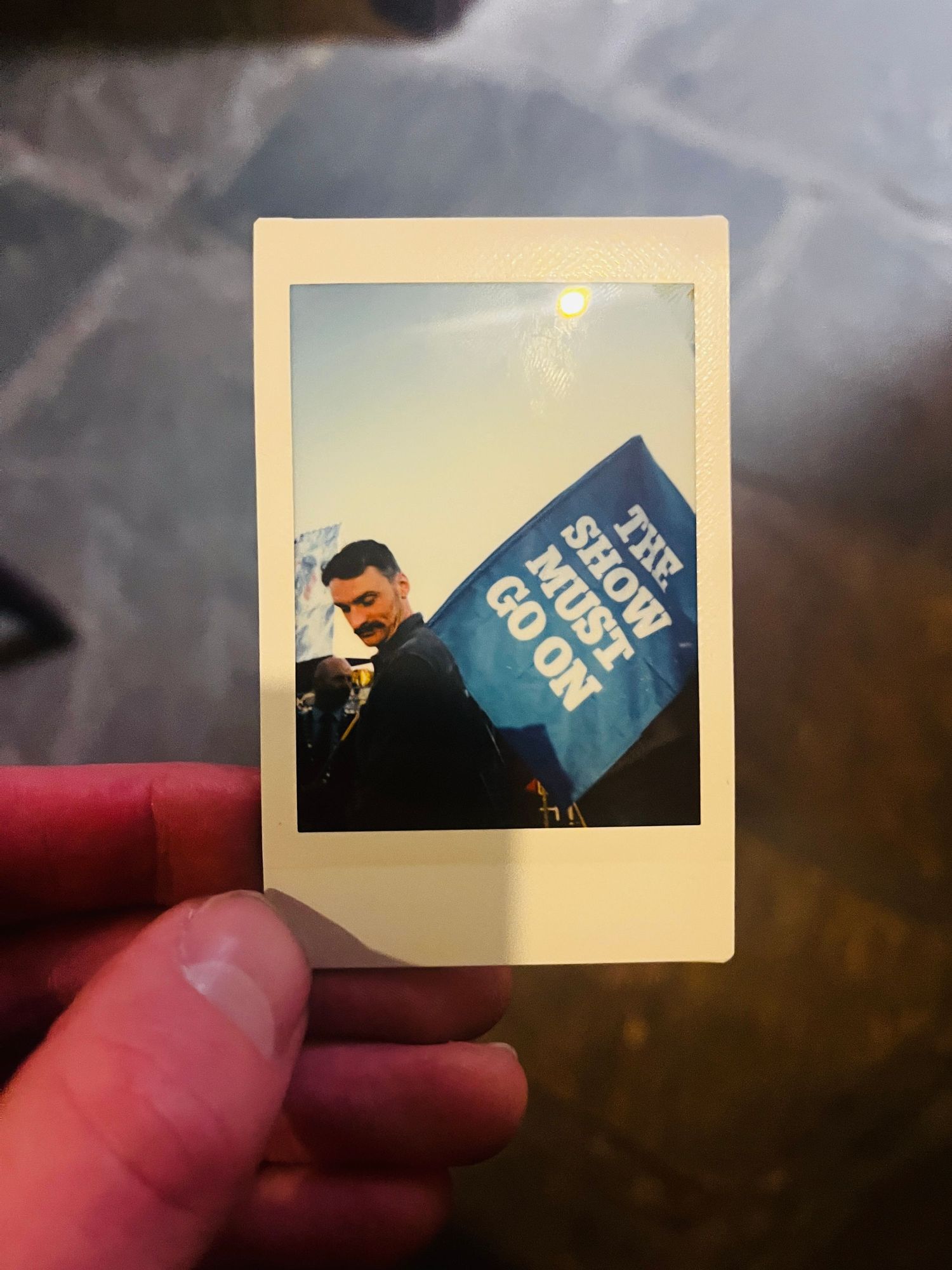 A polaroid photo shows someone holding a blue campaign flag against a bright sky background.