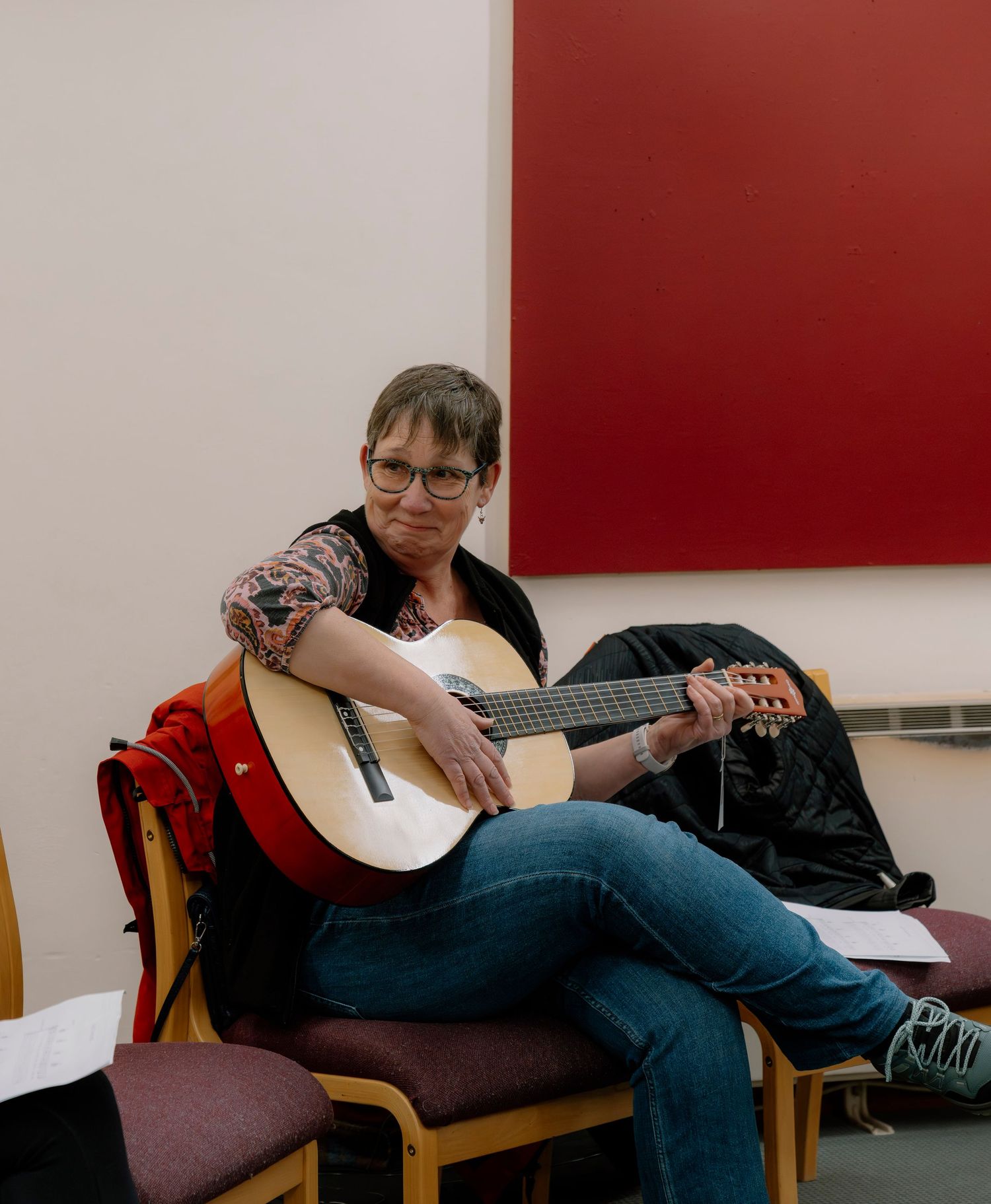Woman with glasses playing an acoustic guitar while seated in a chair with a red painting behind her.