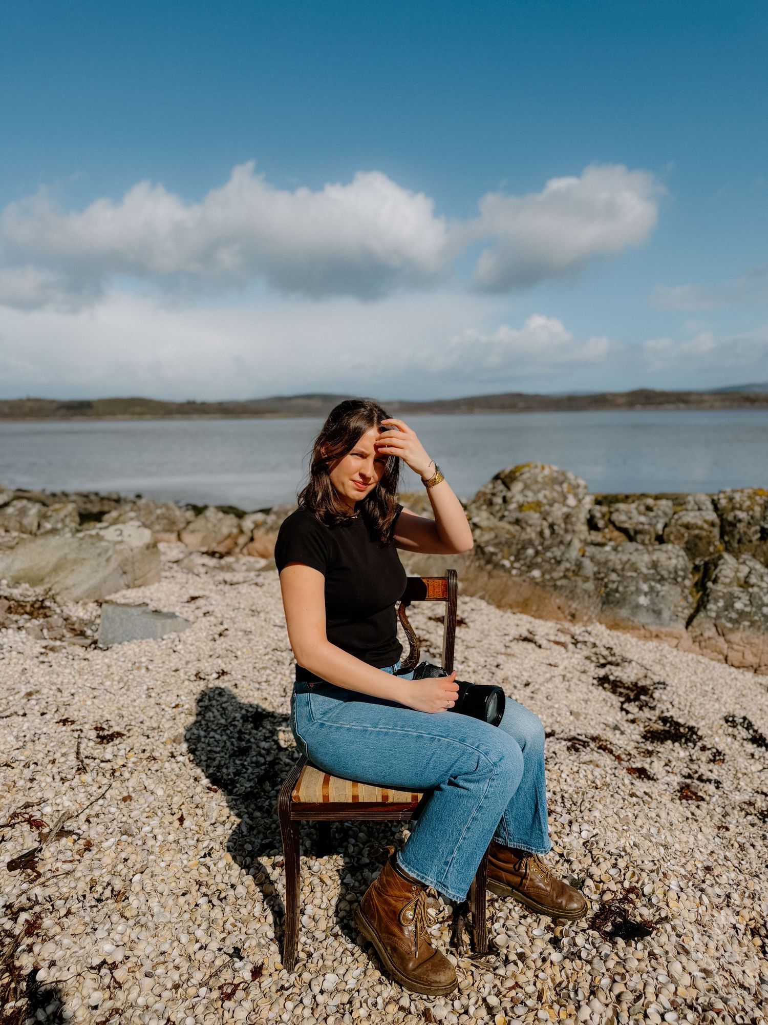 Someone sits on a wooden chair on rocky shoreline with blue water and cloudy sky in background.