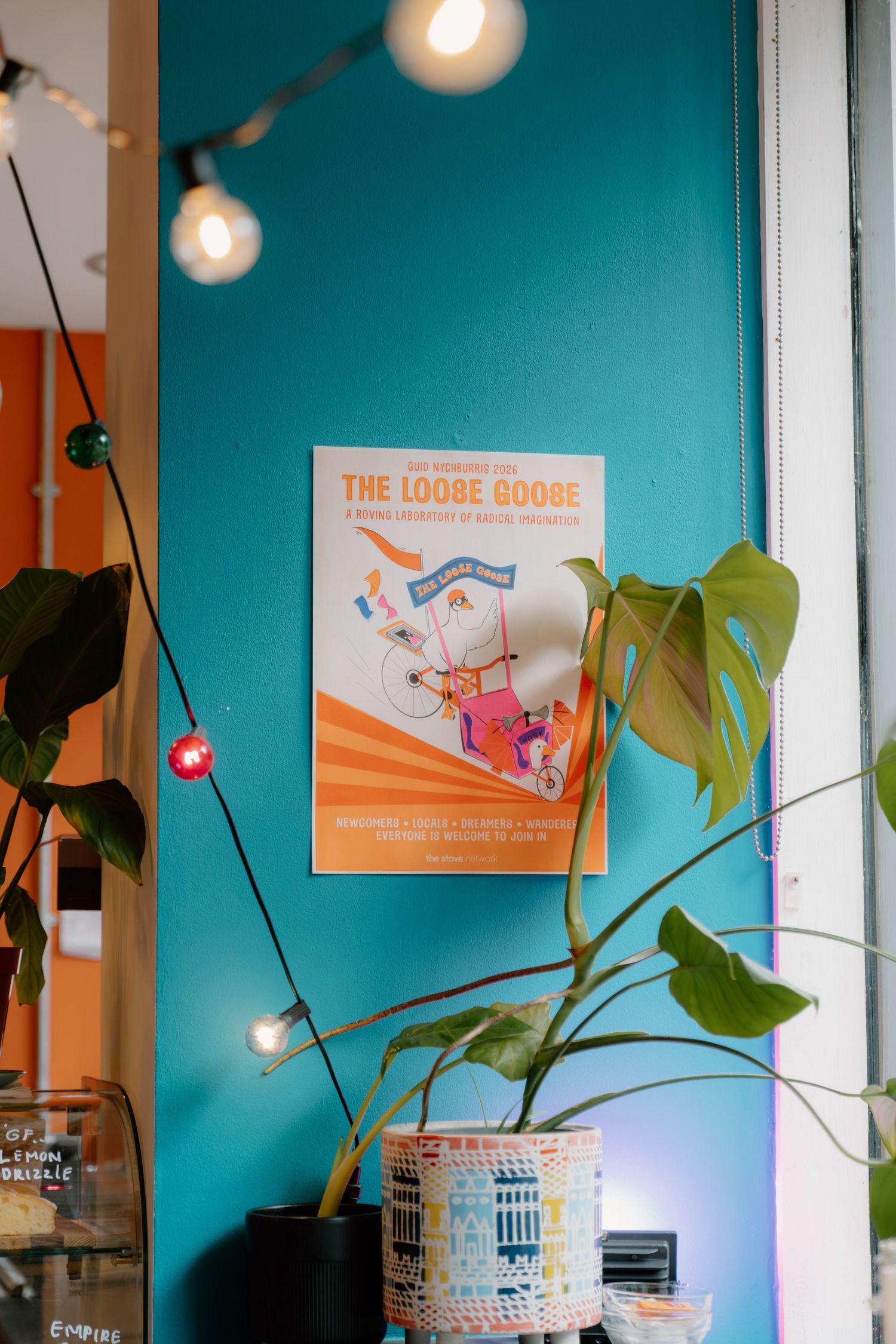Colorful cafe wall with teal paint, The Loose Goose poster, string lights, and a monstera plant in a patterned pot.