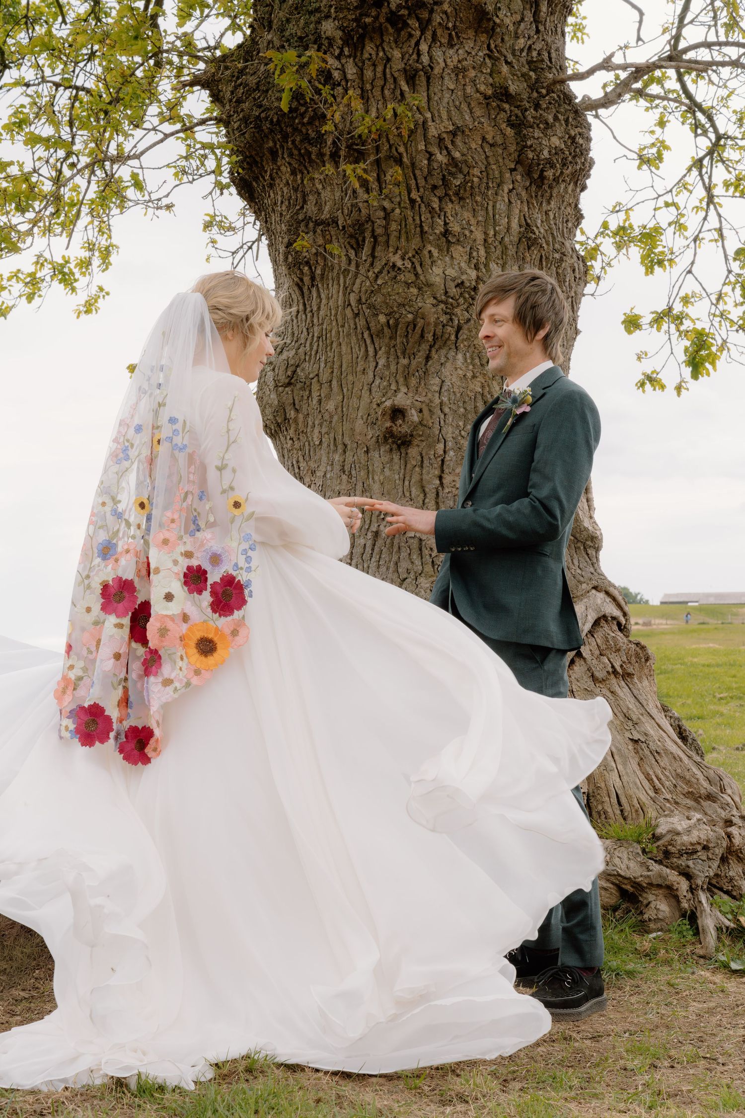 Bride in floral veil and white gown holds groom's hands near large tree outdoors.