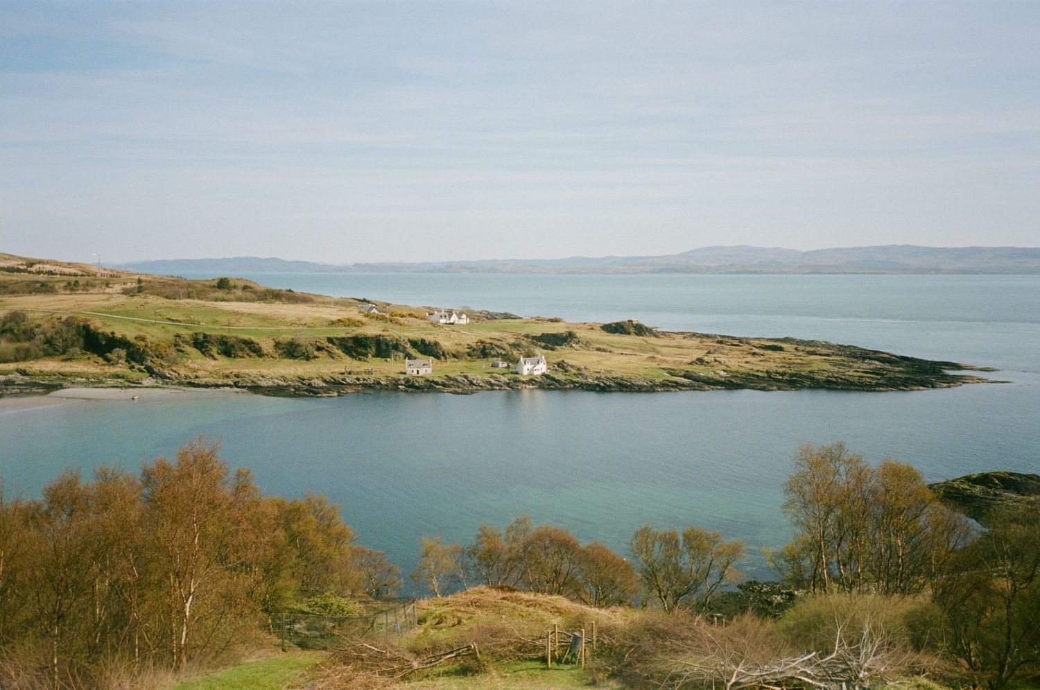 Scenic coastal inlet with turquoise waters, green hills, white cottage, and distant mountains under a clear blue sky.