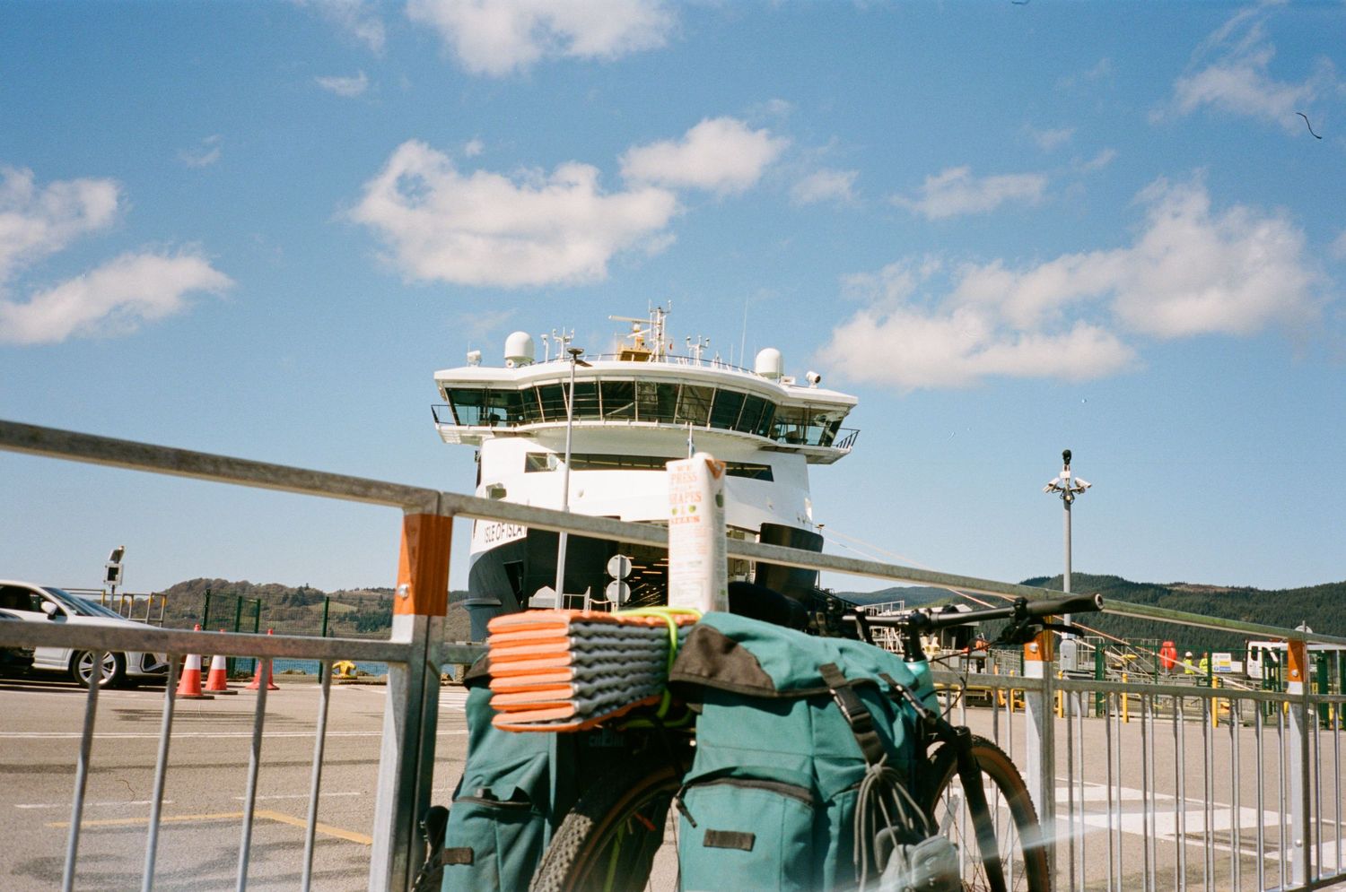 A loaded touring bicycle parked at a ferry terminal with a large white ferry vessel docked under a blue sky.