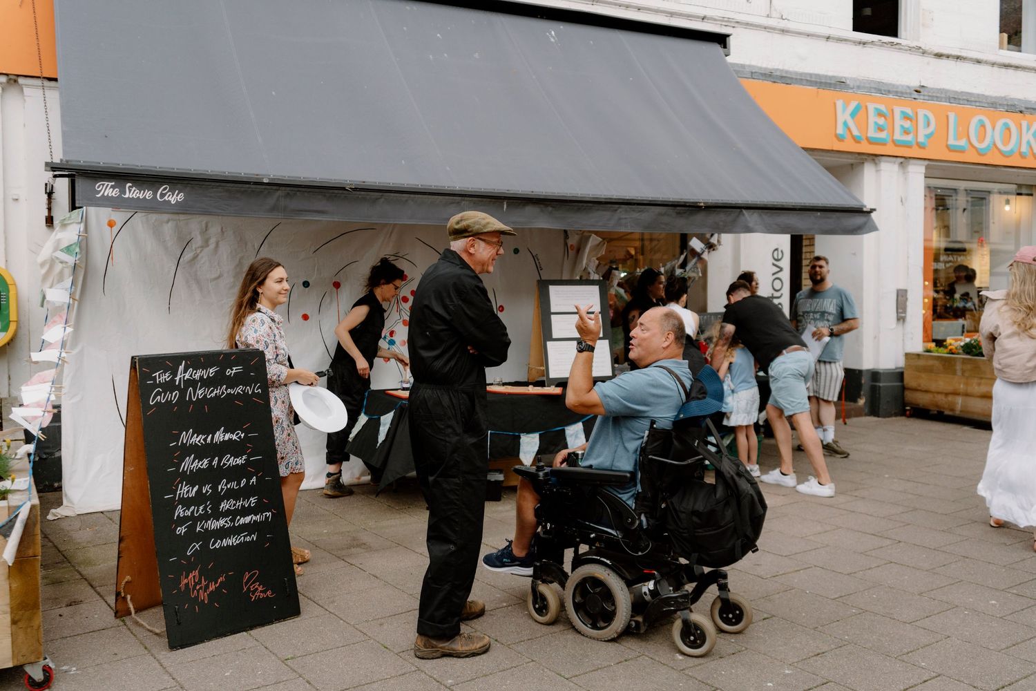 Man in wheelchair conversing outside a cafe with a chalkboard sign, pedestrians and shops visible in background.