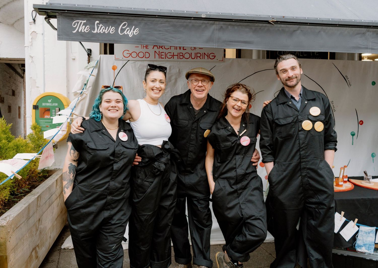 Five people in black jumpsuits smile outside The Stove Cafe, promoting Good Neighbouring community event.
