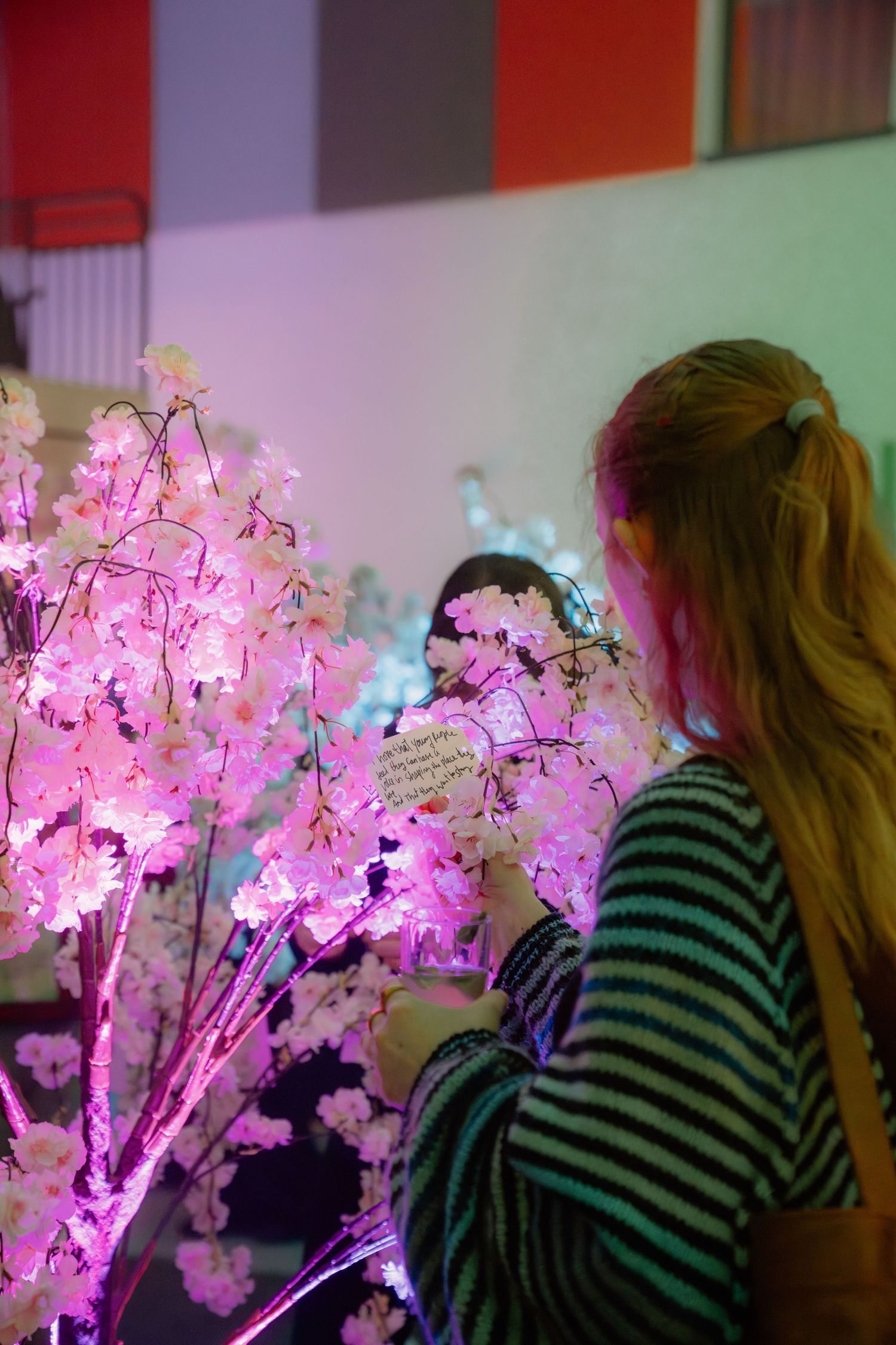 Woman in striped shirt admiring illuminated pink cherry blossom trees with notes attached at an indoor display.