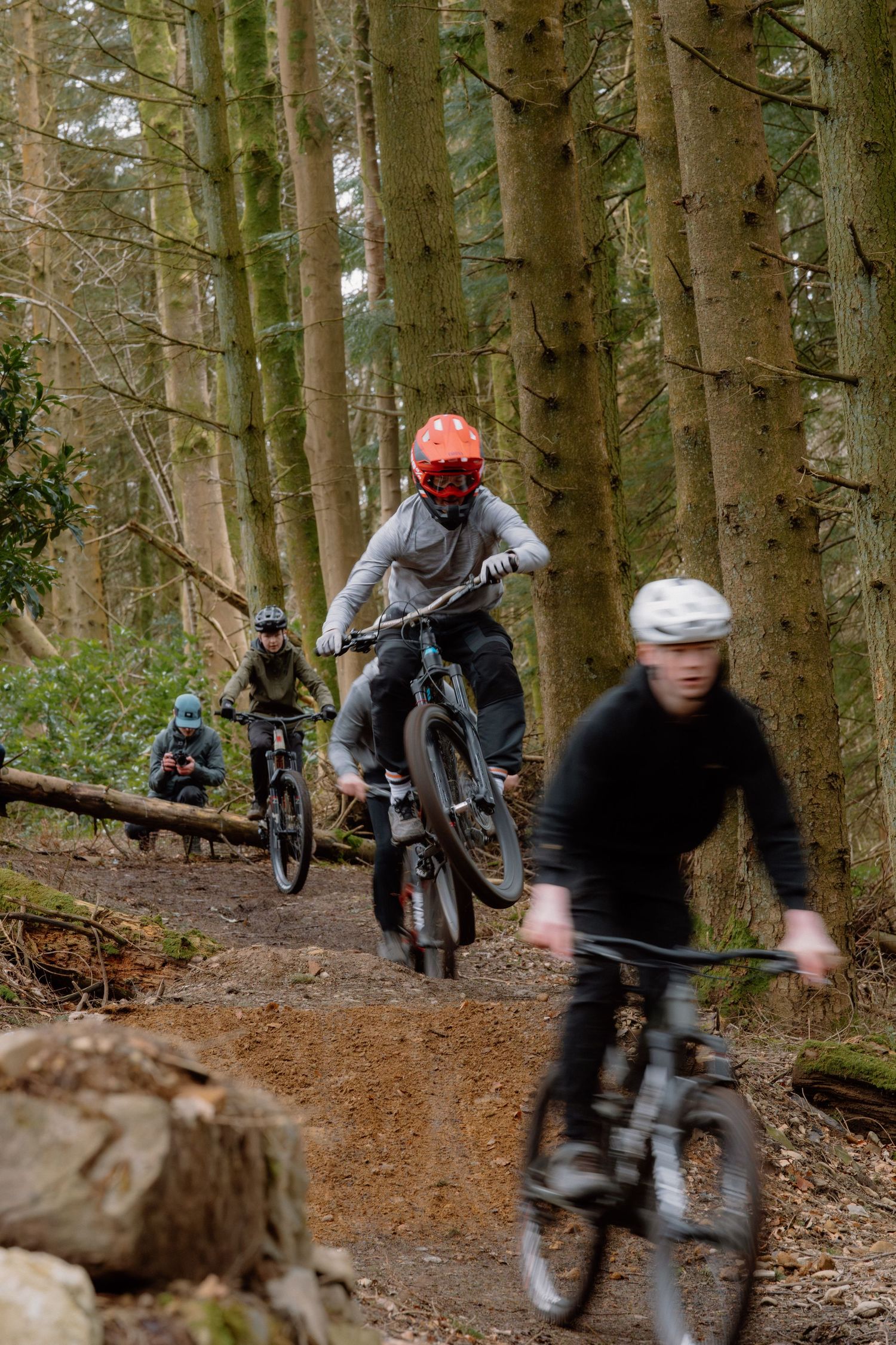 Mountain bikers riding single track trail through dense forest, wearing helmets and protective gear on an outdoor cycling adventure.