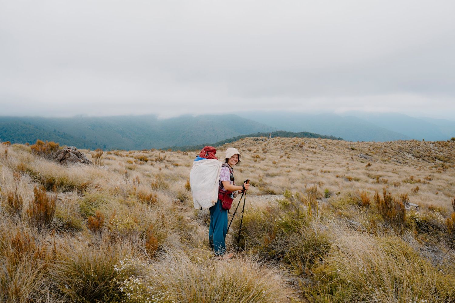People hiking along a coastal trail through tall grass and sand dunes overlooking the ocean on a cloudy day.