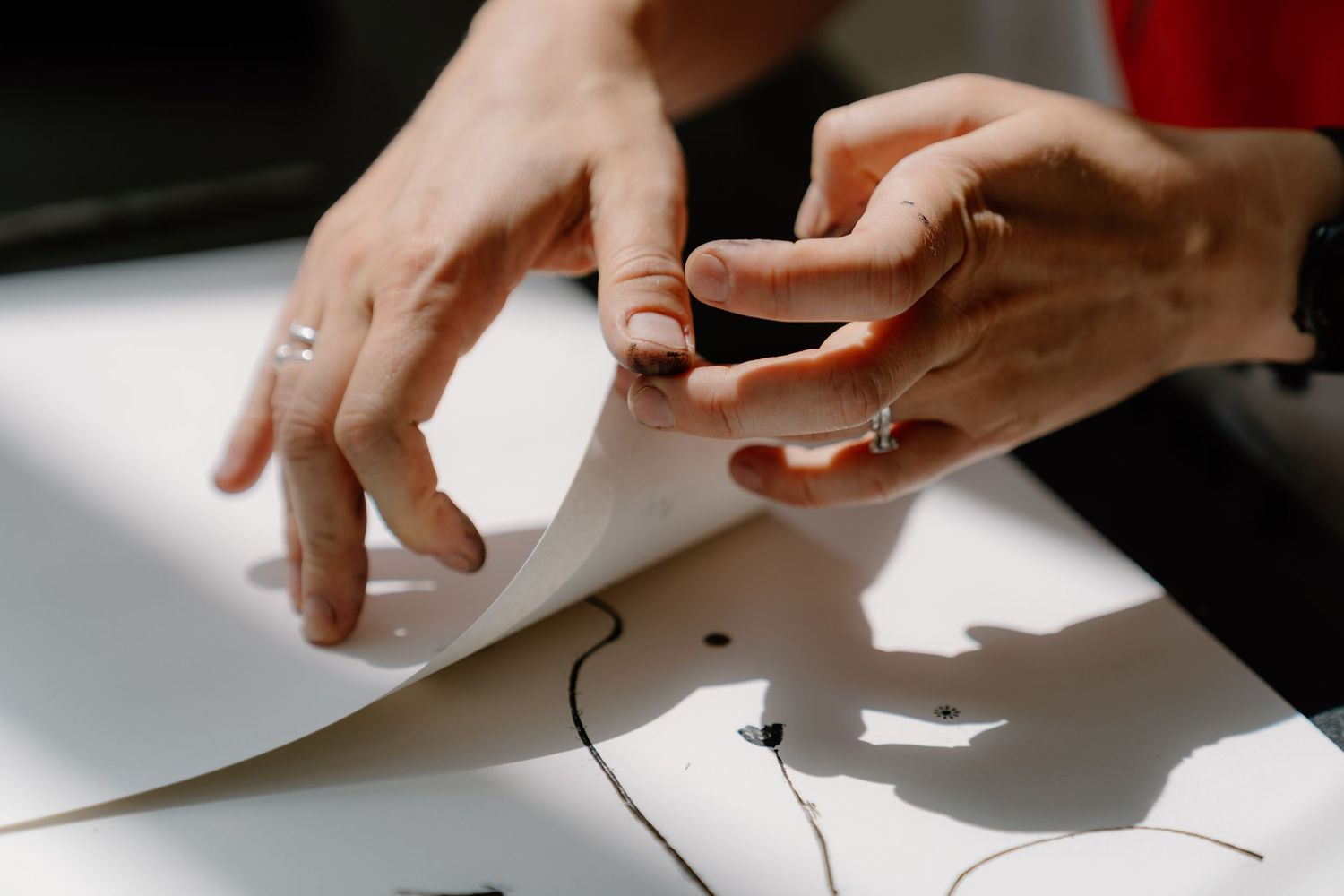 Hands with rings peeling a white paper sheet off a surface with a black line design visible underneath.