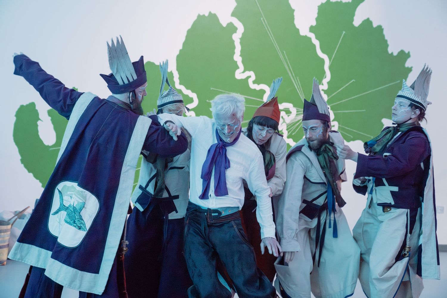Group of performers in colorful costumes posing dramatically against a green painted backdrop.
