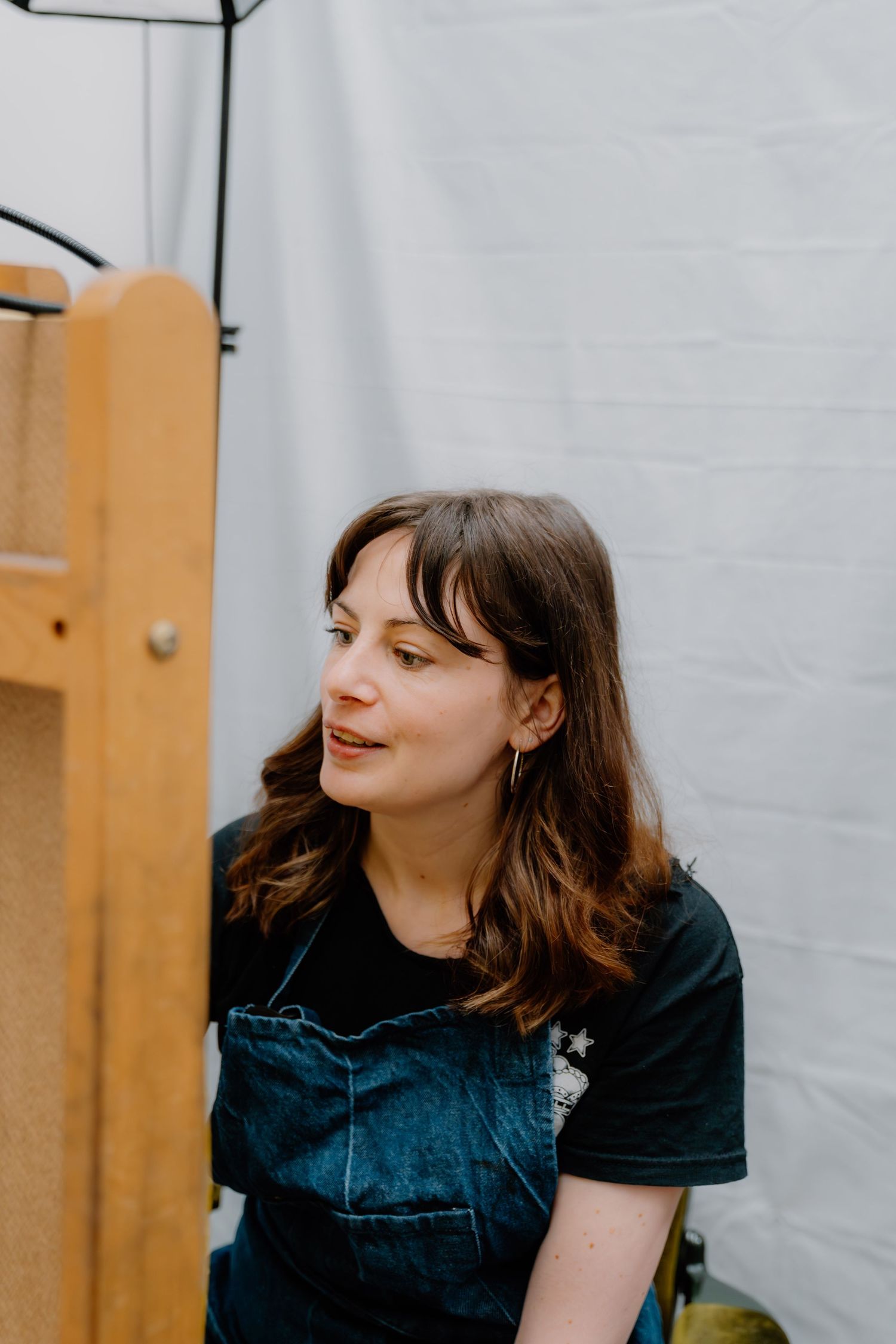 A young woman with brown hair wearing a denim apron over a black t-shirt, looking to the side in a bright studio.