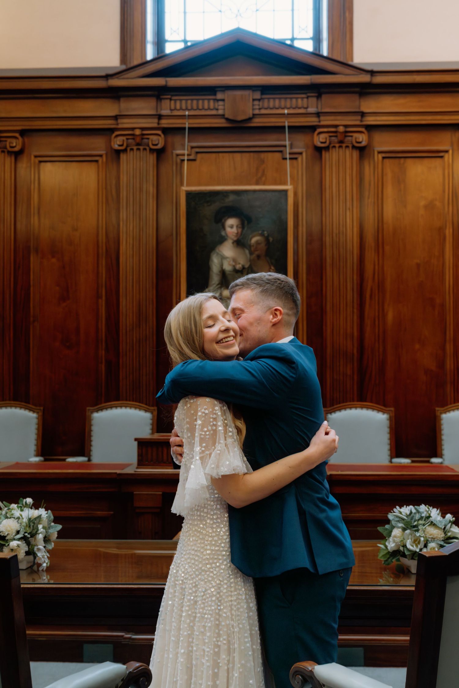 Bride and groom share a joyful embrace in an elegant wood-paneled wedding ceremony room with floral arrangements.