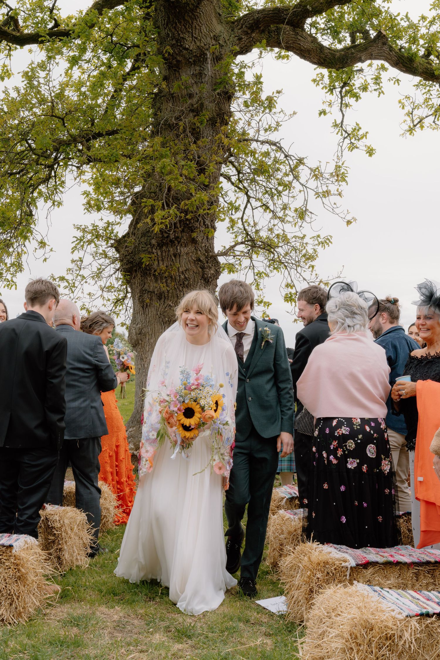 A bride and groom walk down an outdoor aisle lined with hay bales under a large oak tree at a rustic countryside wedding.
