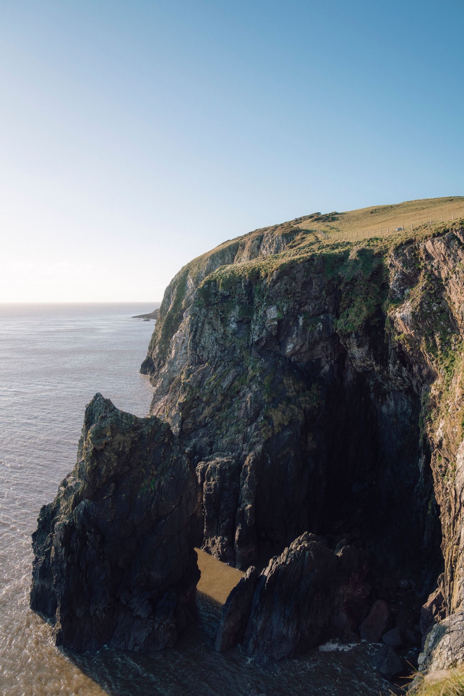 Dramatic aerial view of rugged coastal cliffs rising above the ocean with sunlight illuminating the rocky landscape.