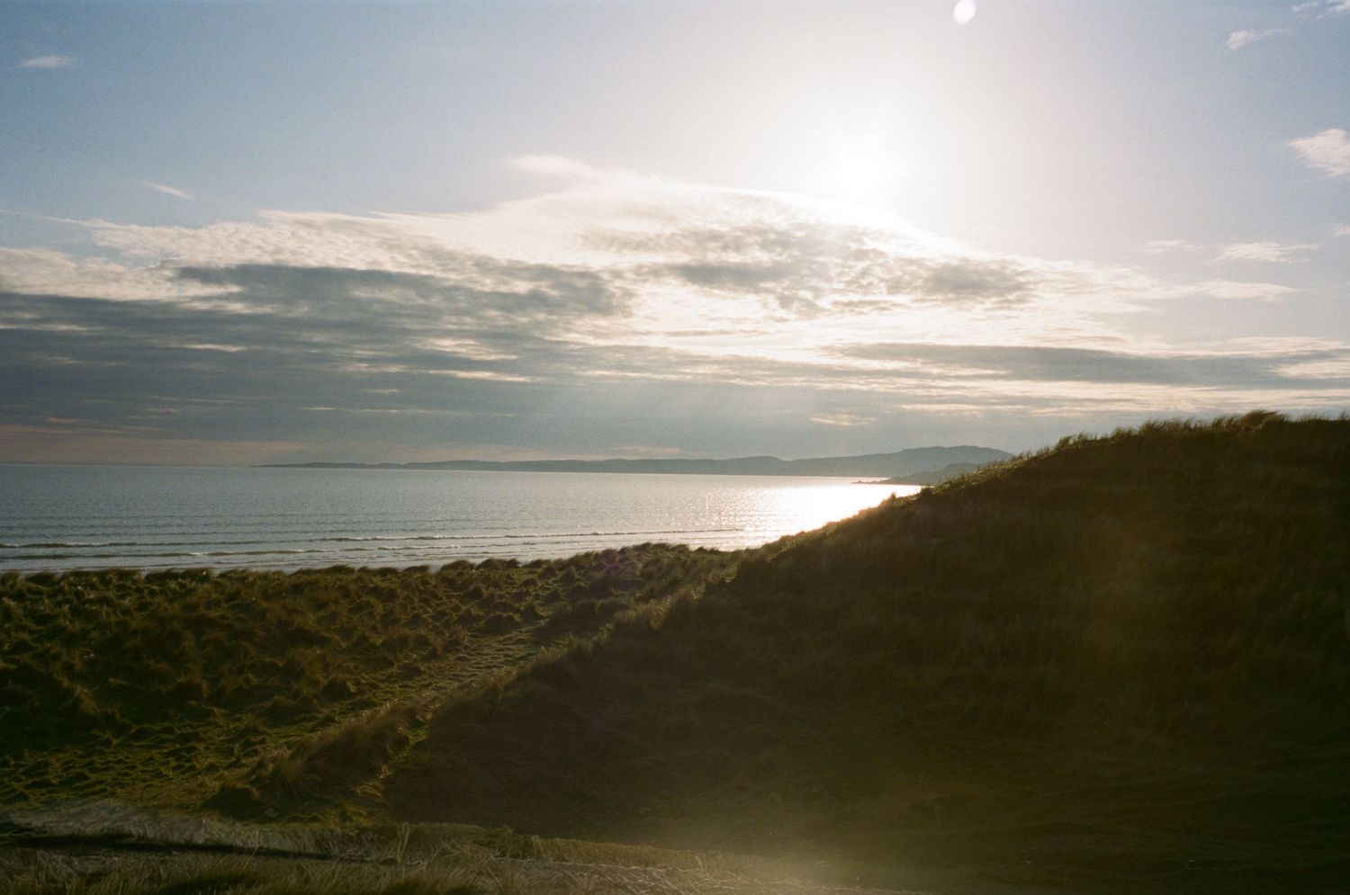Sunlit coastal dunes with green grass overlooking a calm sea under a partly cloudy sky at golden hour.
