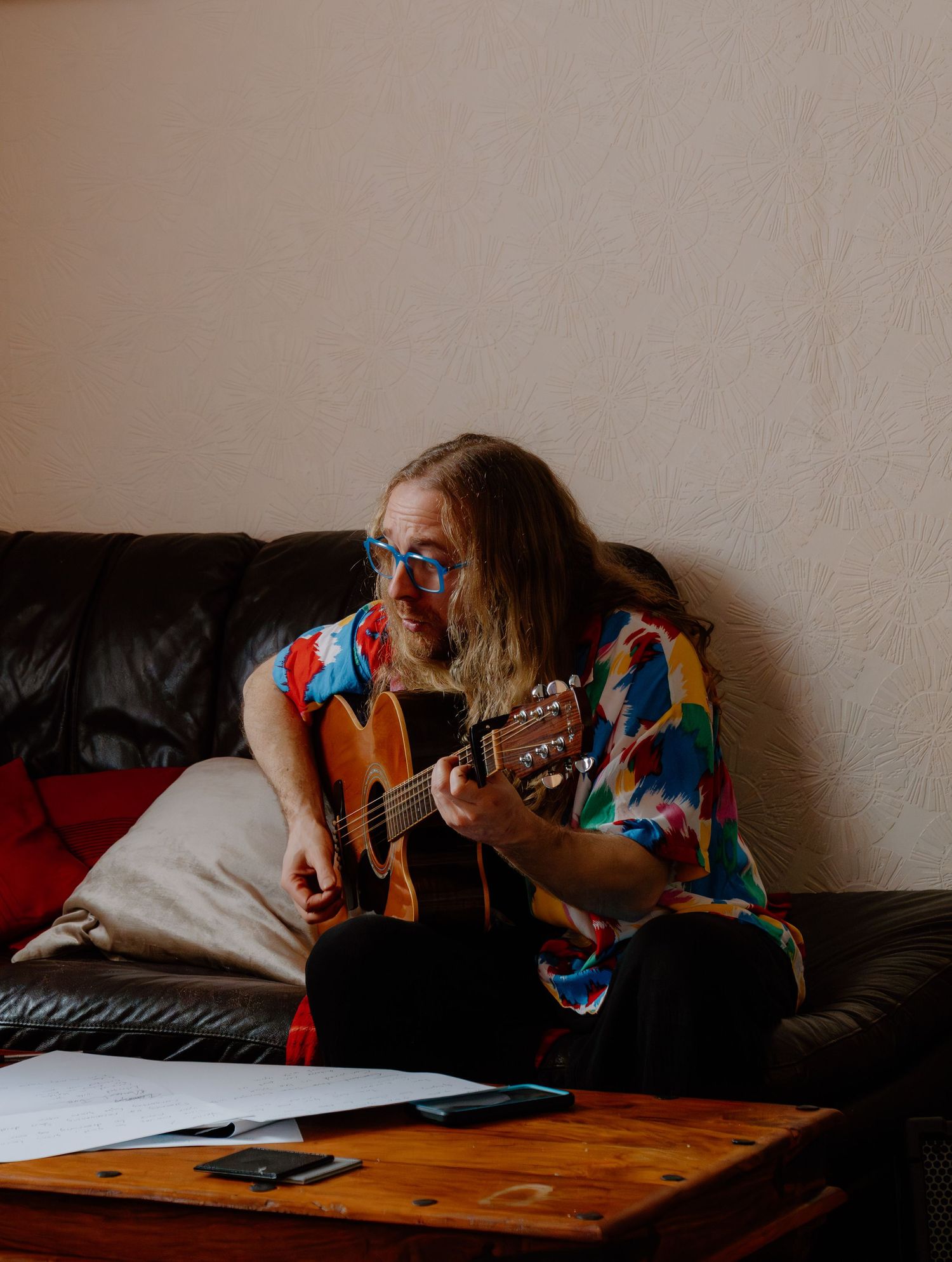 Long-haired musician in colorful floral shirt and blue glasses playing acoustic guitar on black leather sofa.