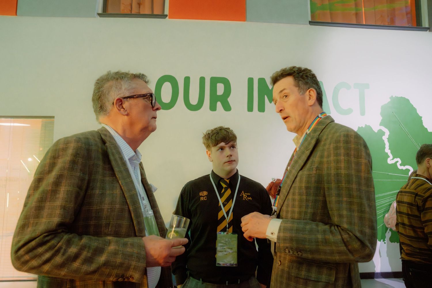 Two men in suits converse at a corporate event, with a young attendee and green branded wall visible behind them.