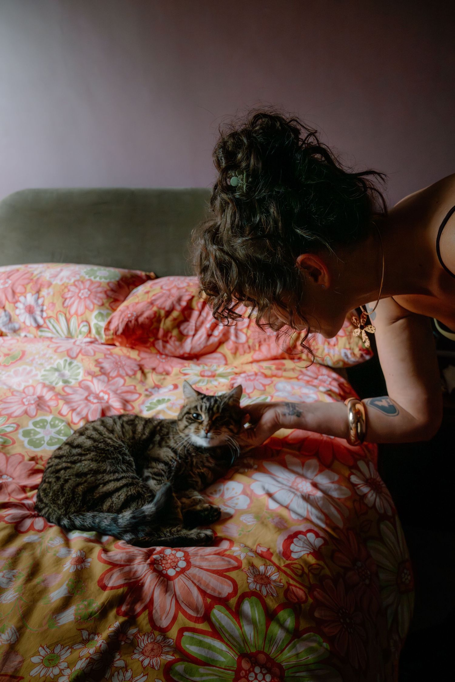 Woman with curly hair petting a tabby cat lying on a floral pink bedspread indoors.