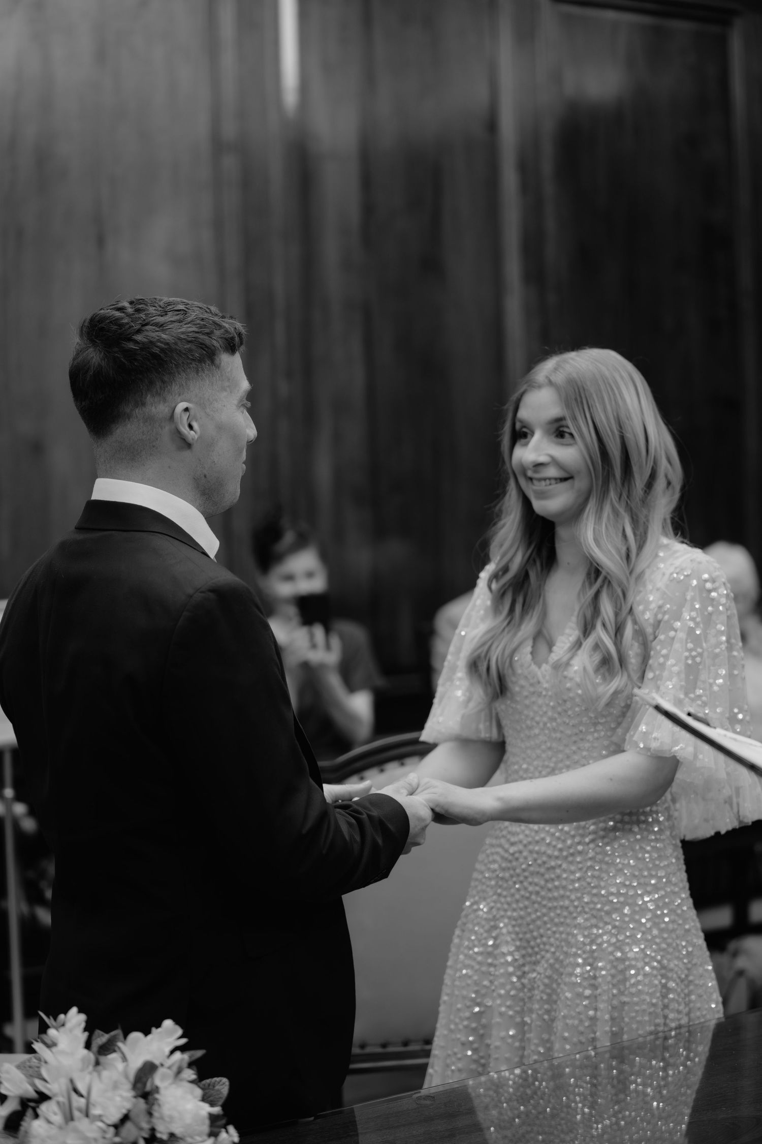 Black and white photo of a smiling bride in a sequined dress holding hands with a groom during a wedding ceremony.