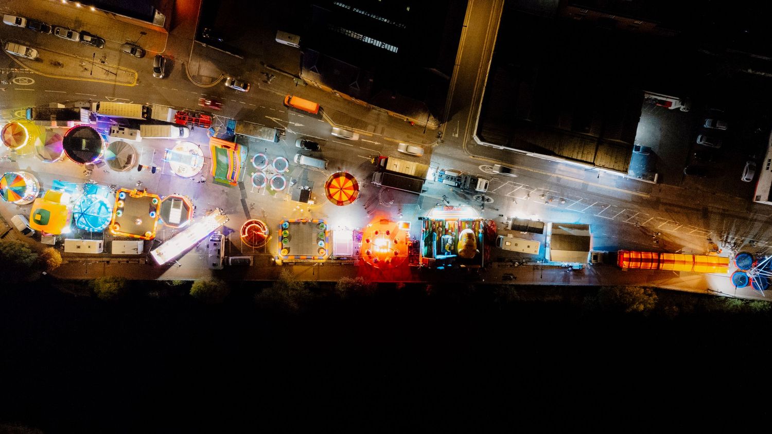 Aerial night view of illuminated shopping center parking lot with cars and bright storefronts.