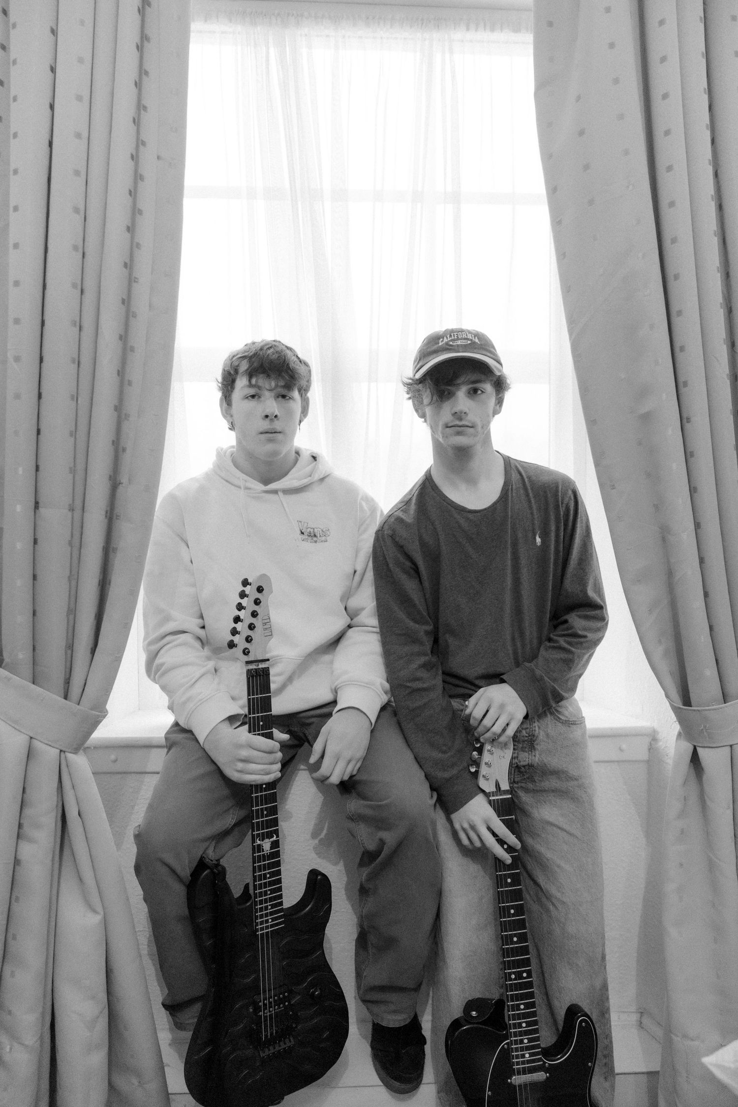 Two young musicians sitting on a windowsill holding electric guitars in a black and white photo.