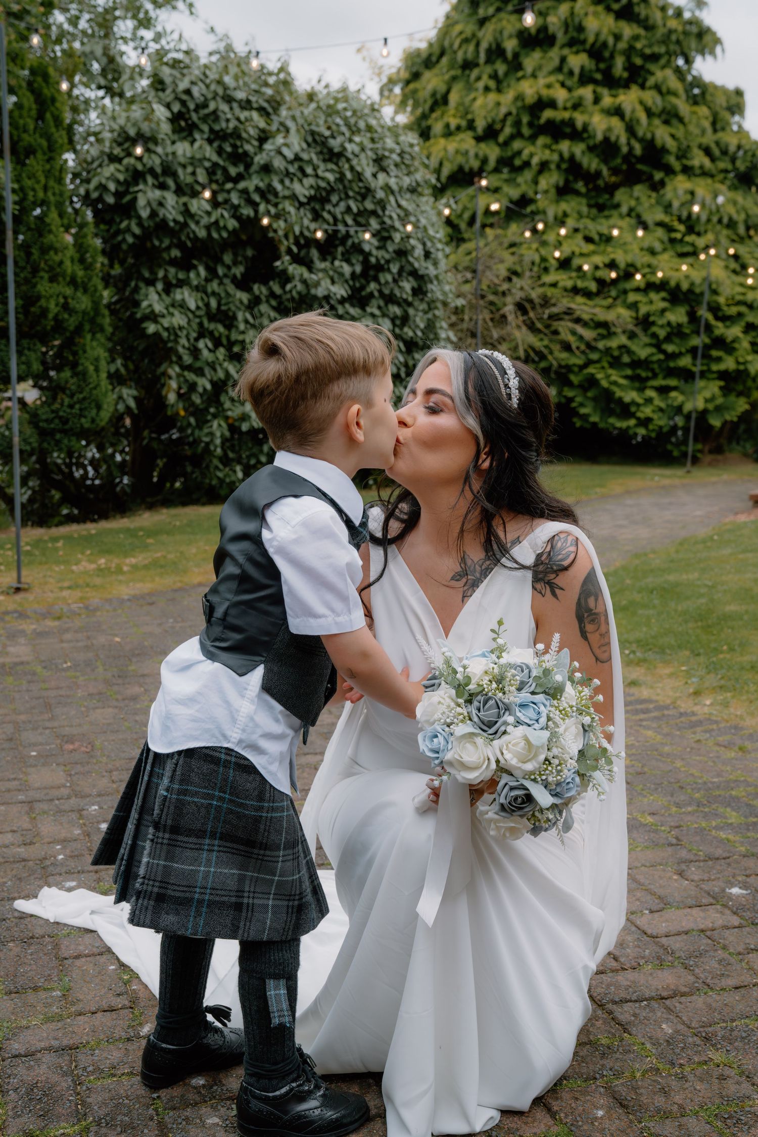 Bride in white gown kisses young boy in tartan kilt while holding blue and white bouquet outdoors.
