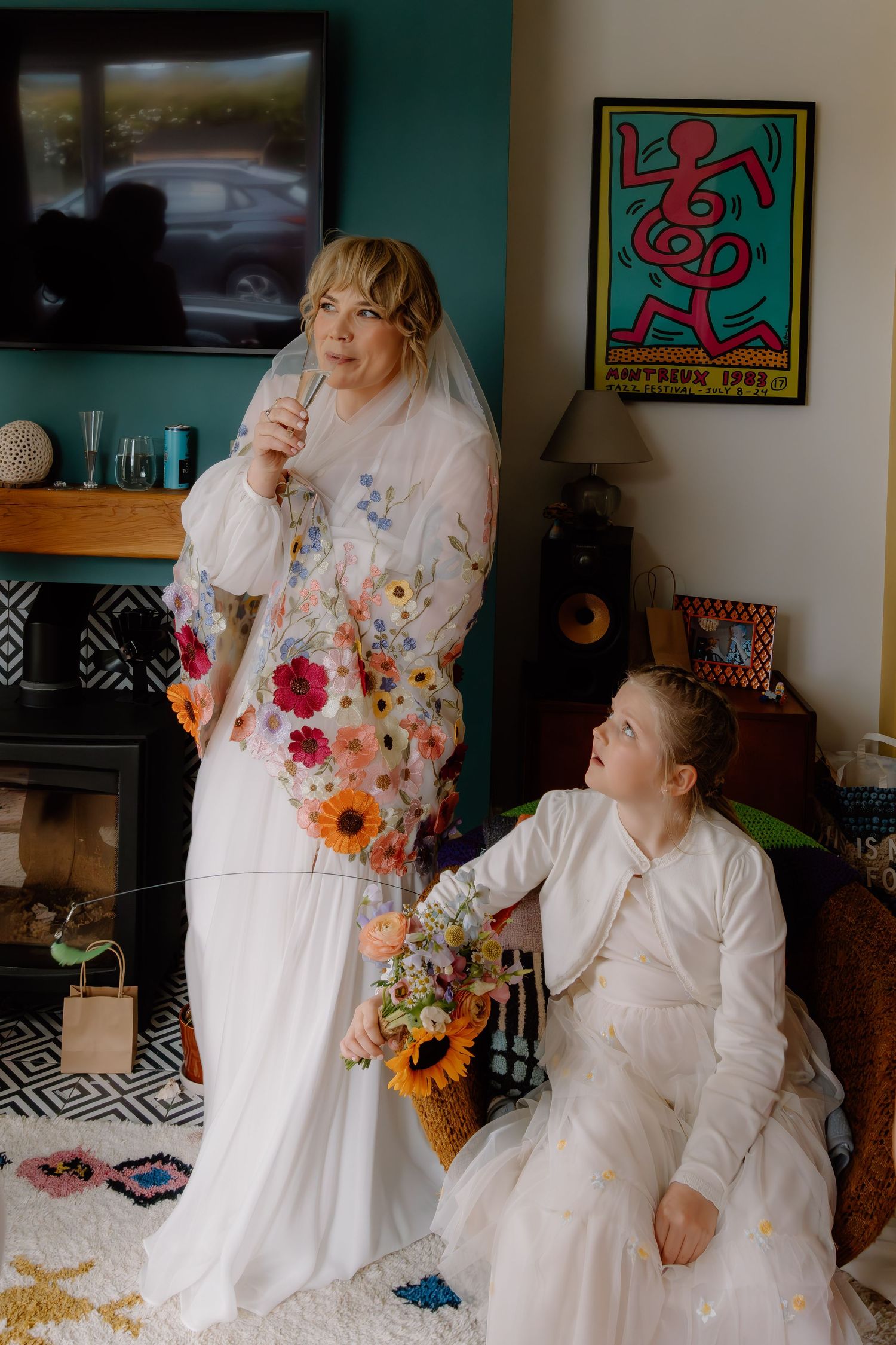 Bride in floral veil holding mic with child in white outfit holding flower basket in colorful decorated room.