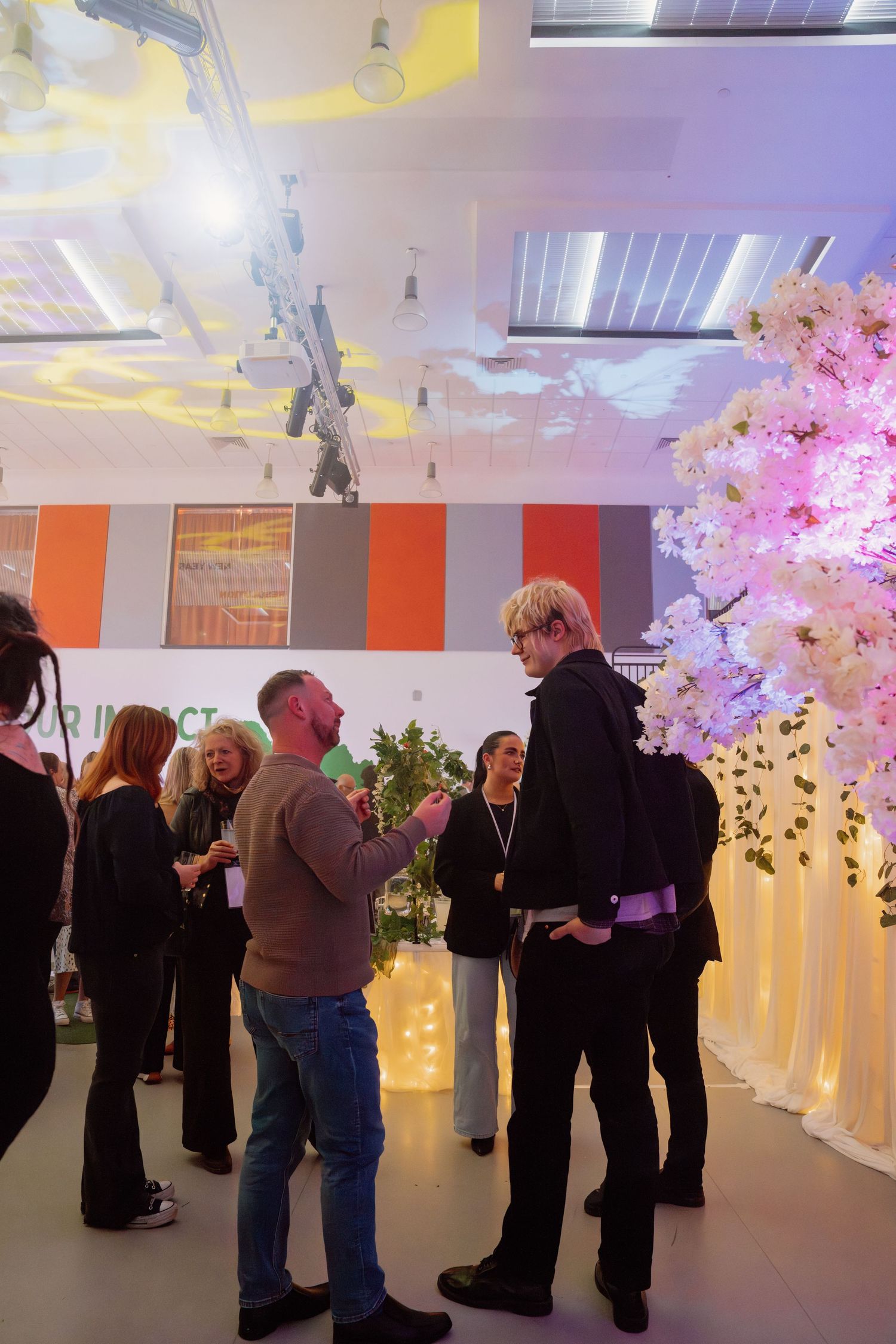 People mingling at an indoor event with pink floral decorations and colorful lighting in a modern venue.