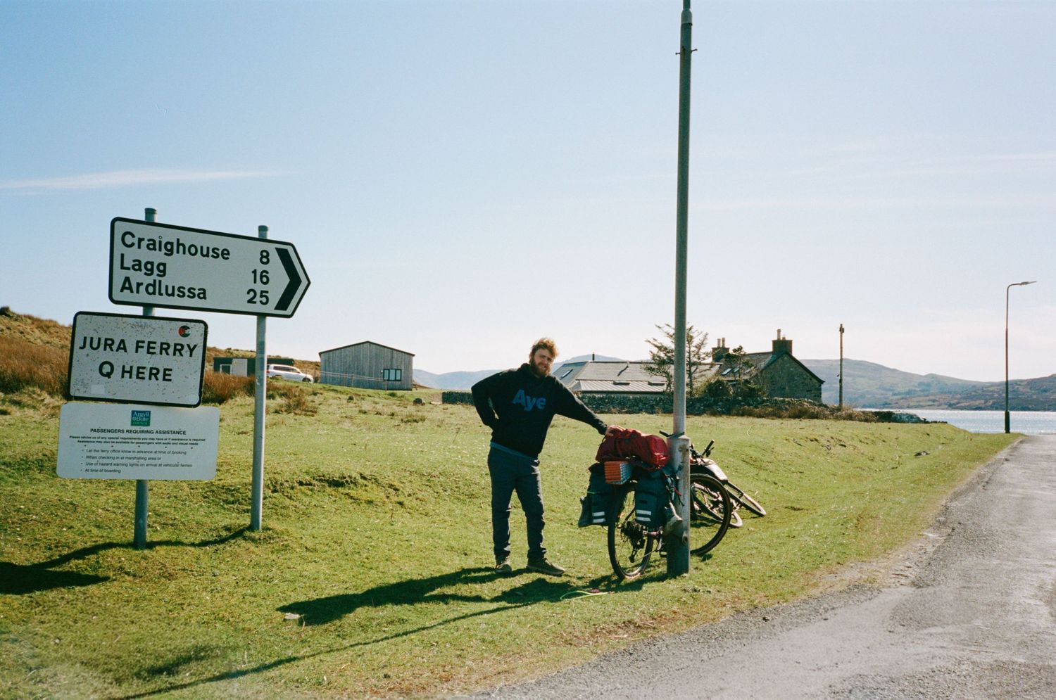 Cyclist with loaded touring bike at Jura Ferry road sign on Scottish island, with loch and cottage in background.