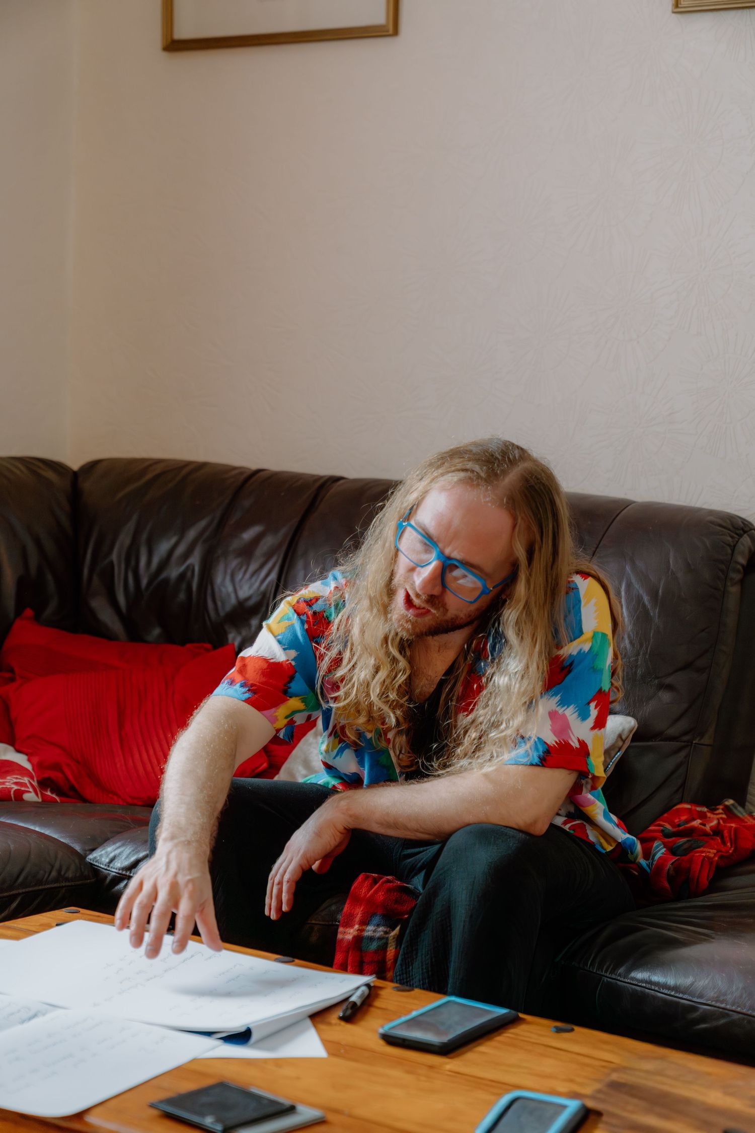 Person with long hair and blue glasses reviewing documents on a leather sofa wearing a colorful floral shirt.