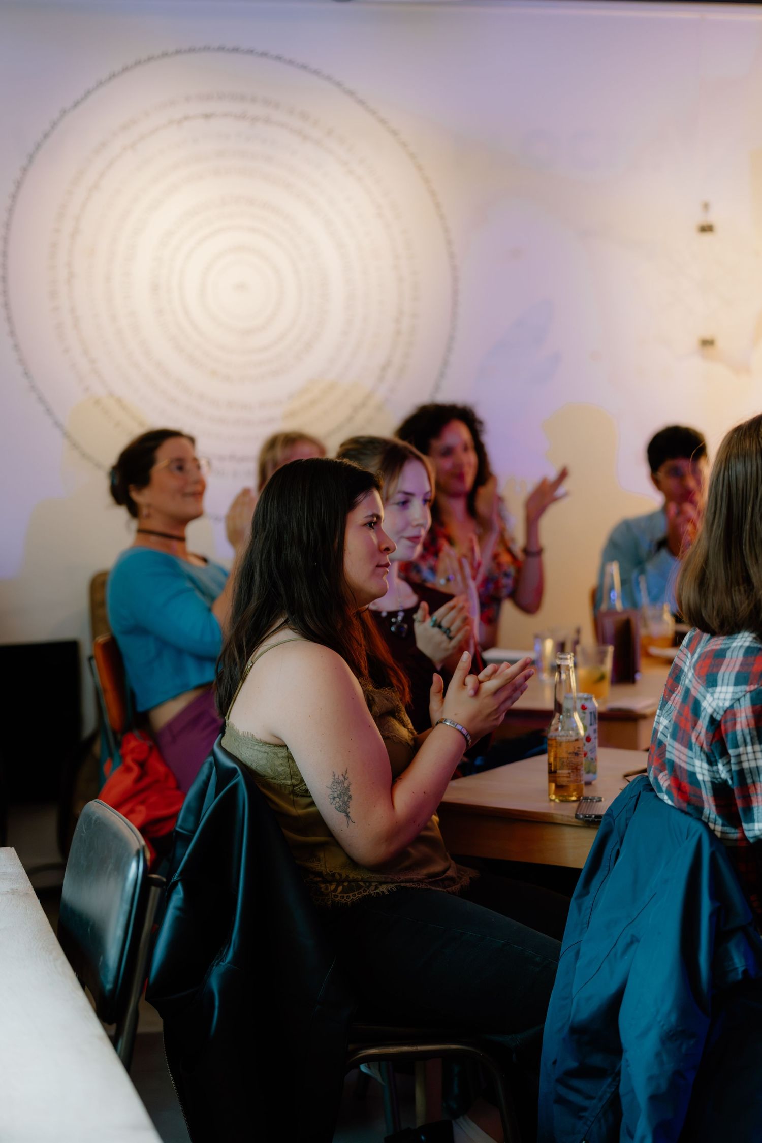 Group of women socializing at a casual indoor gathering, seated at tables with drinks, spiral artwork on wall behind them.