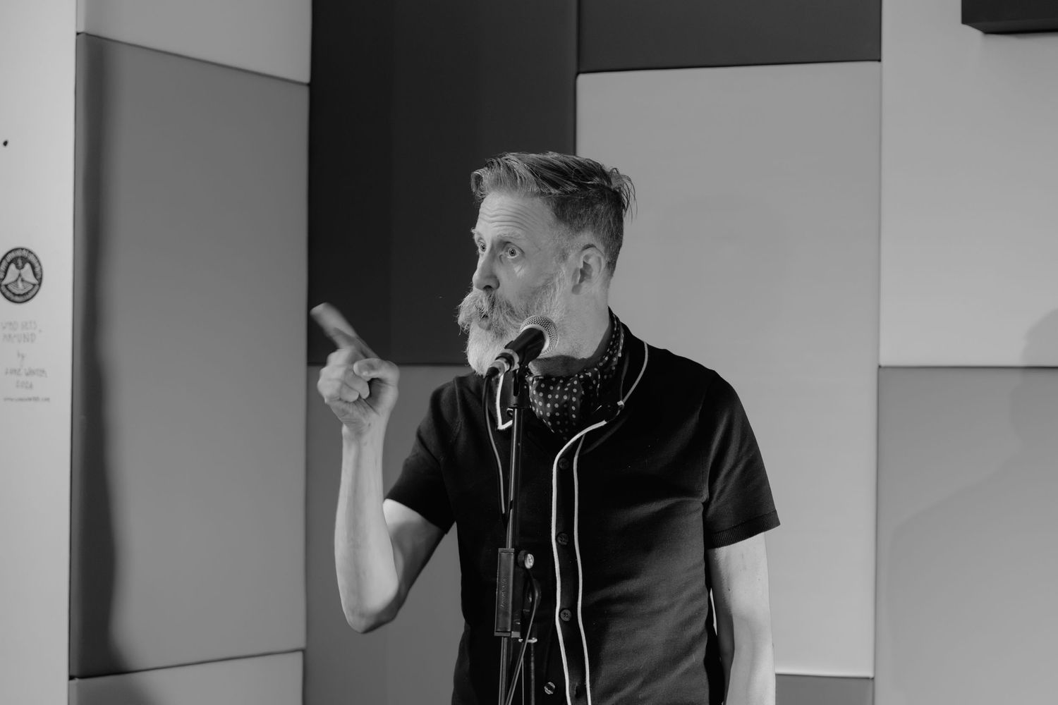Black and white photo of a man pointing while speaking, wearing a black shirt with headphones around his neck.