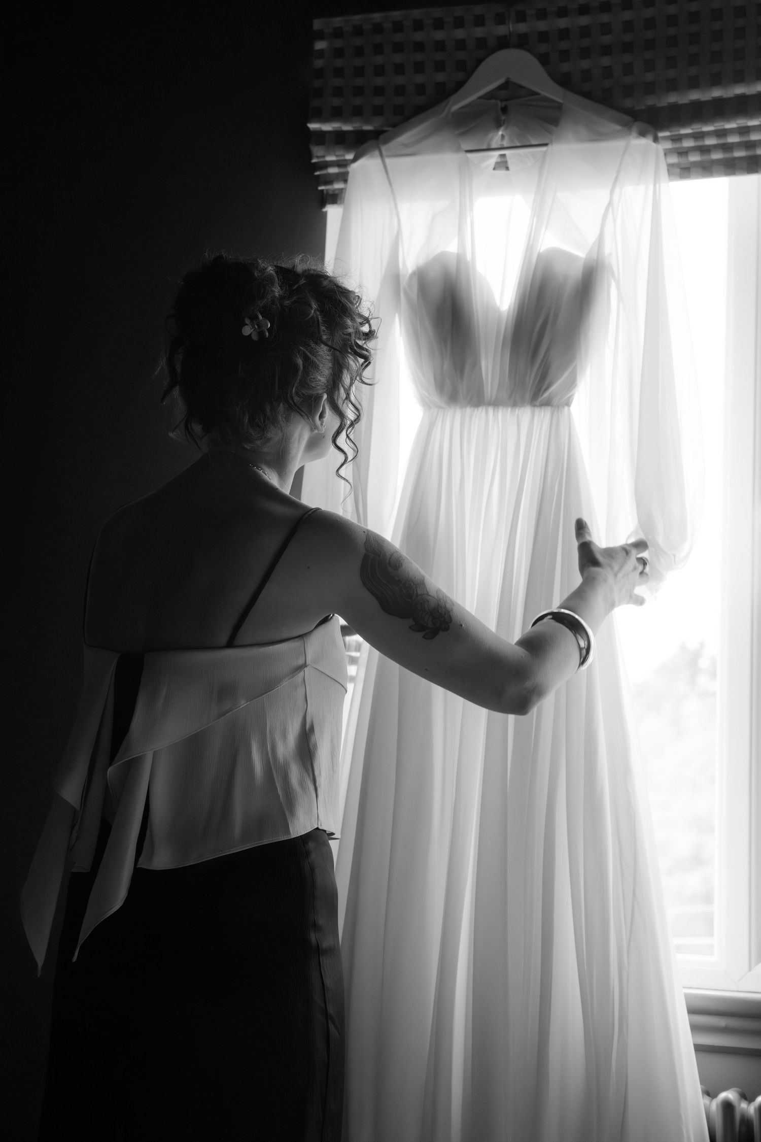 Black and white photo of bride reaching for elegant wedding gown hanging by window before ceremony.