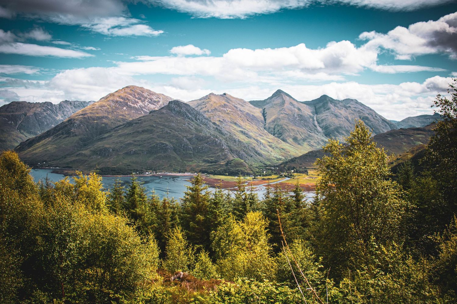 Panoramic view of snow-capped mountains and lakes in the Scottish Highlands with dramatic clouds and golden sunlight.