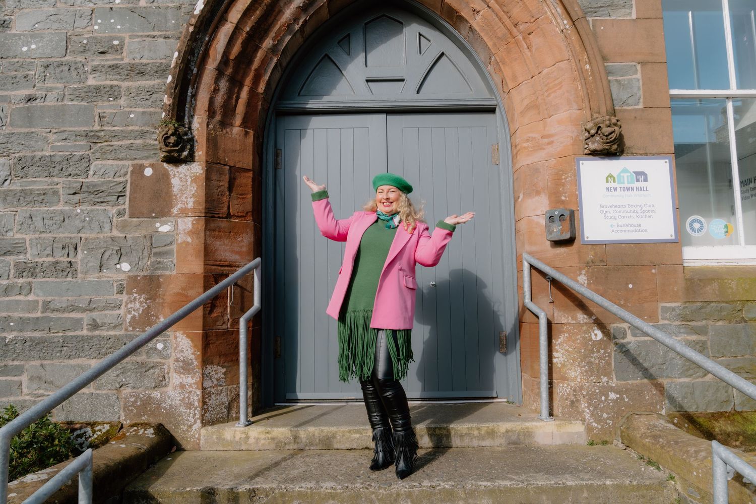 Woman in pink coat and green beret poses at stone church entrance with grey doors on sunny day.