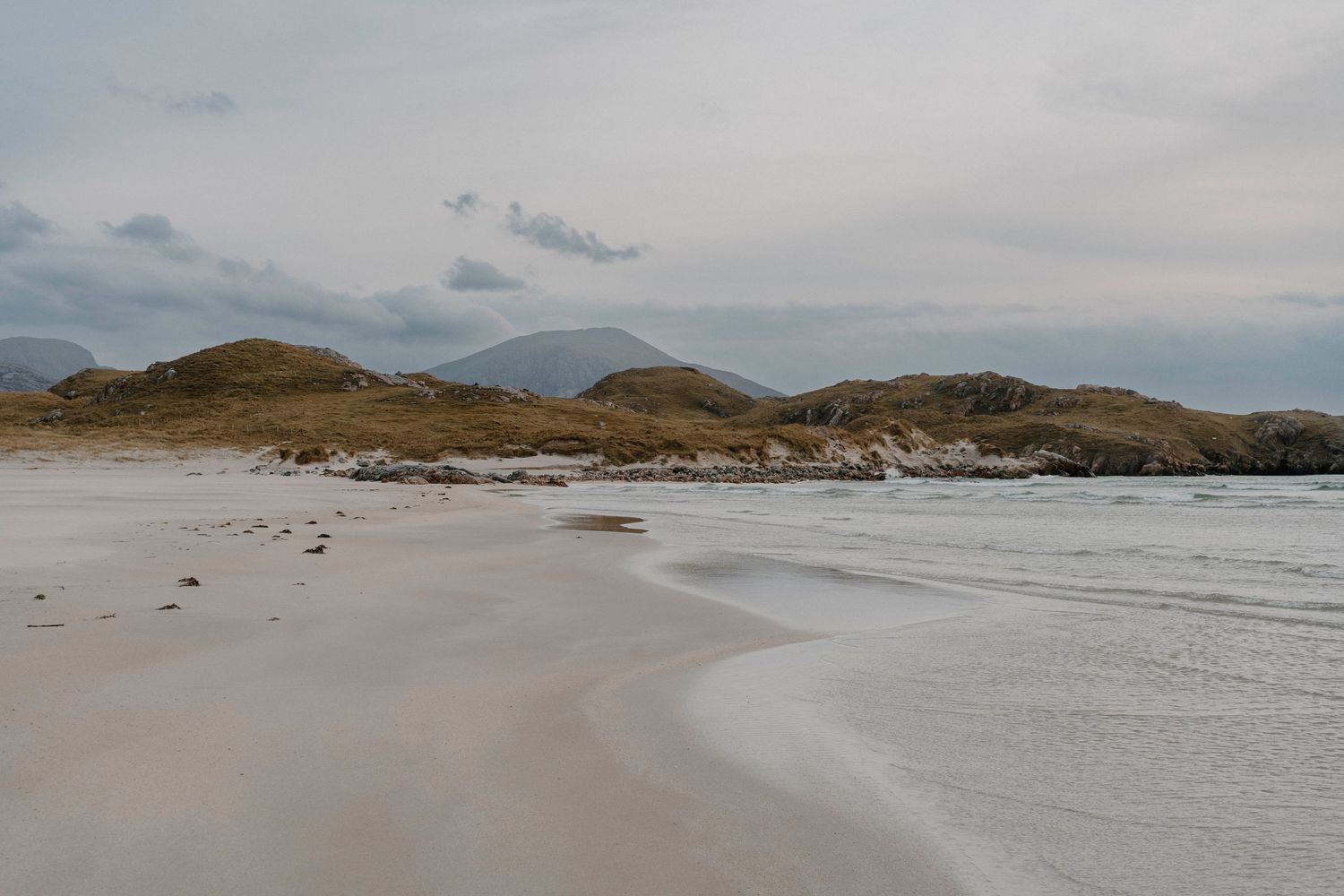 A serene sandy beach with gentle waves, rolling green hills, and misty mountains under a cloudy sky.