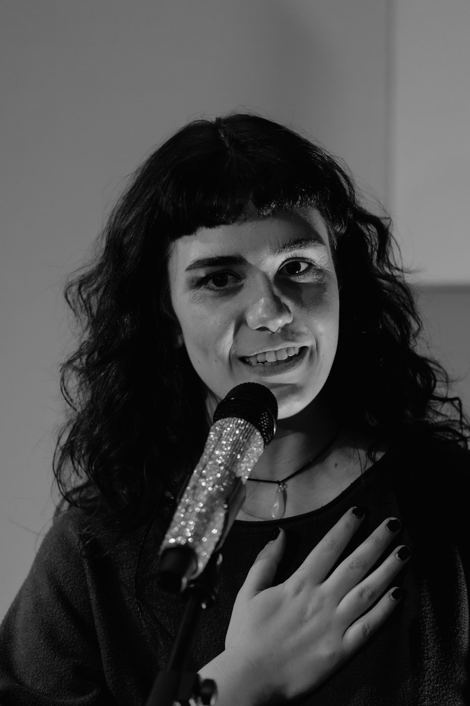 Black and white portrait of a woman with curly dark hair speaking into a microphone with hand on chest.