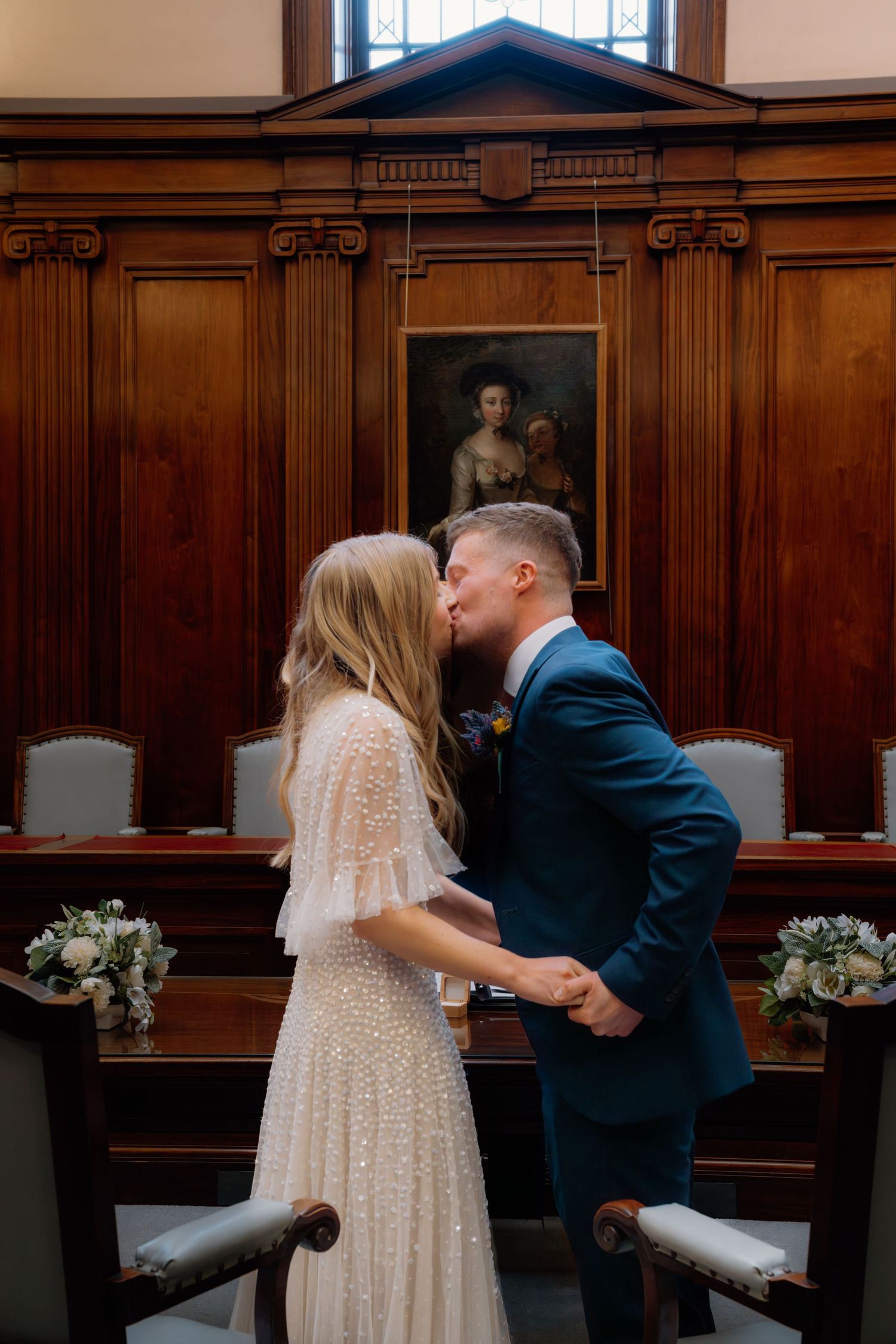 A couple shares their first kiss as newlyweds in an elegant wood-paneled ceremony room with floral arrangements nearby.