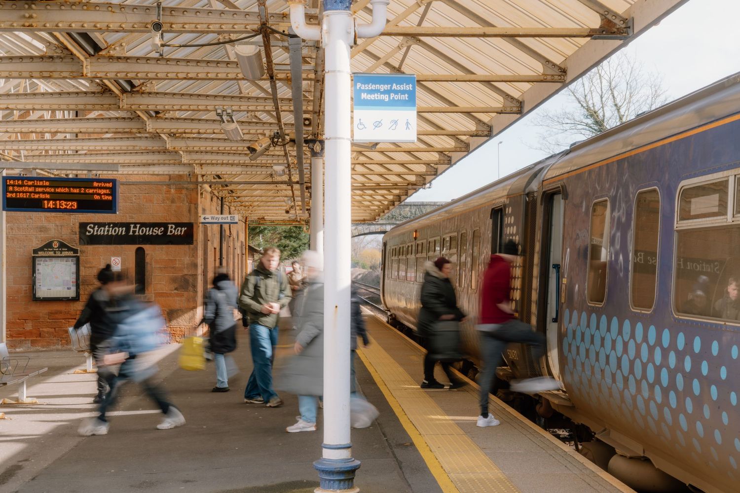 Passengers boarding a train at a busy UK railway station platform with a Station House Bar sign visible.