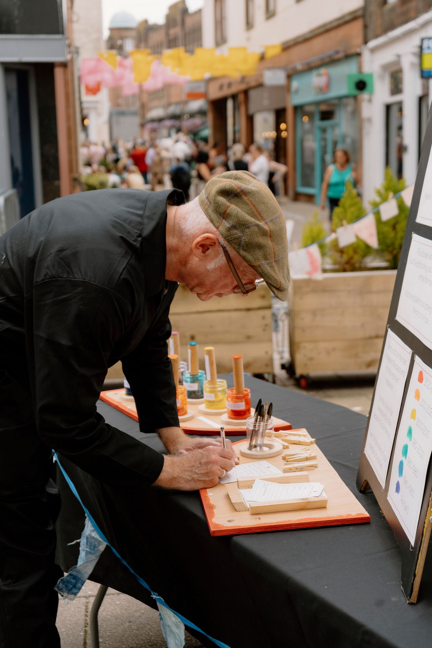 Elderly man in black jacket and flat cap carefully writing at an outdoor market stall with colorful paint tools.