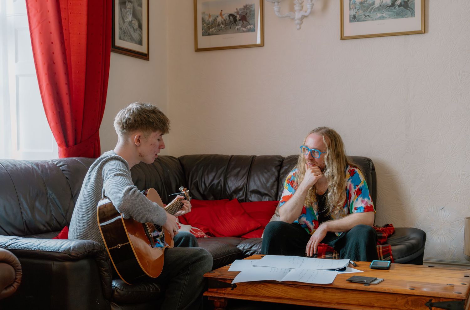Young man playing acoustic guitar for a girl on a leather sofa in a cozy living room.