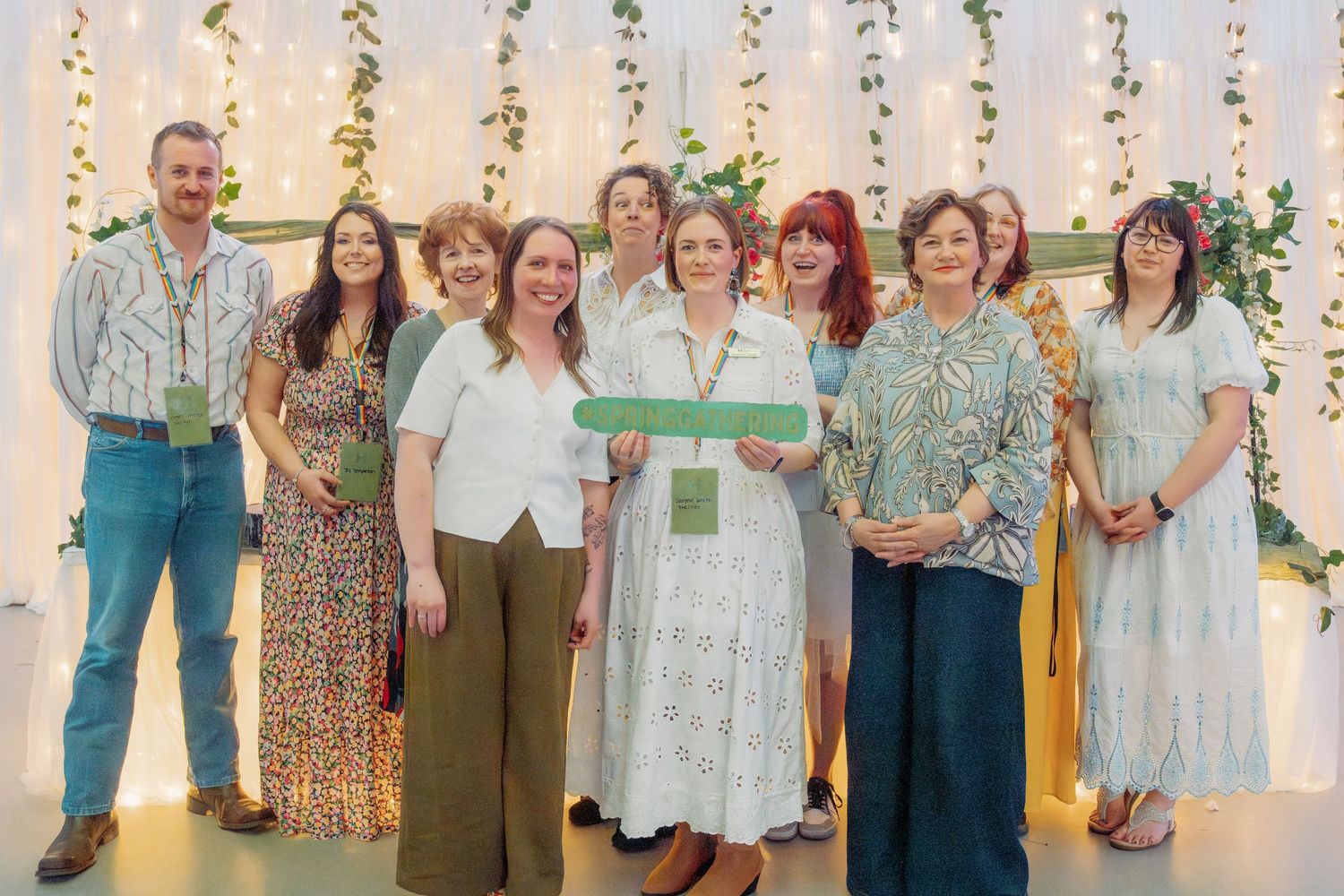 Group of nine people posing together at an elegant event with floral backdrop and string lights.