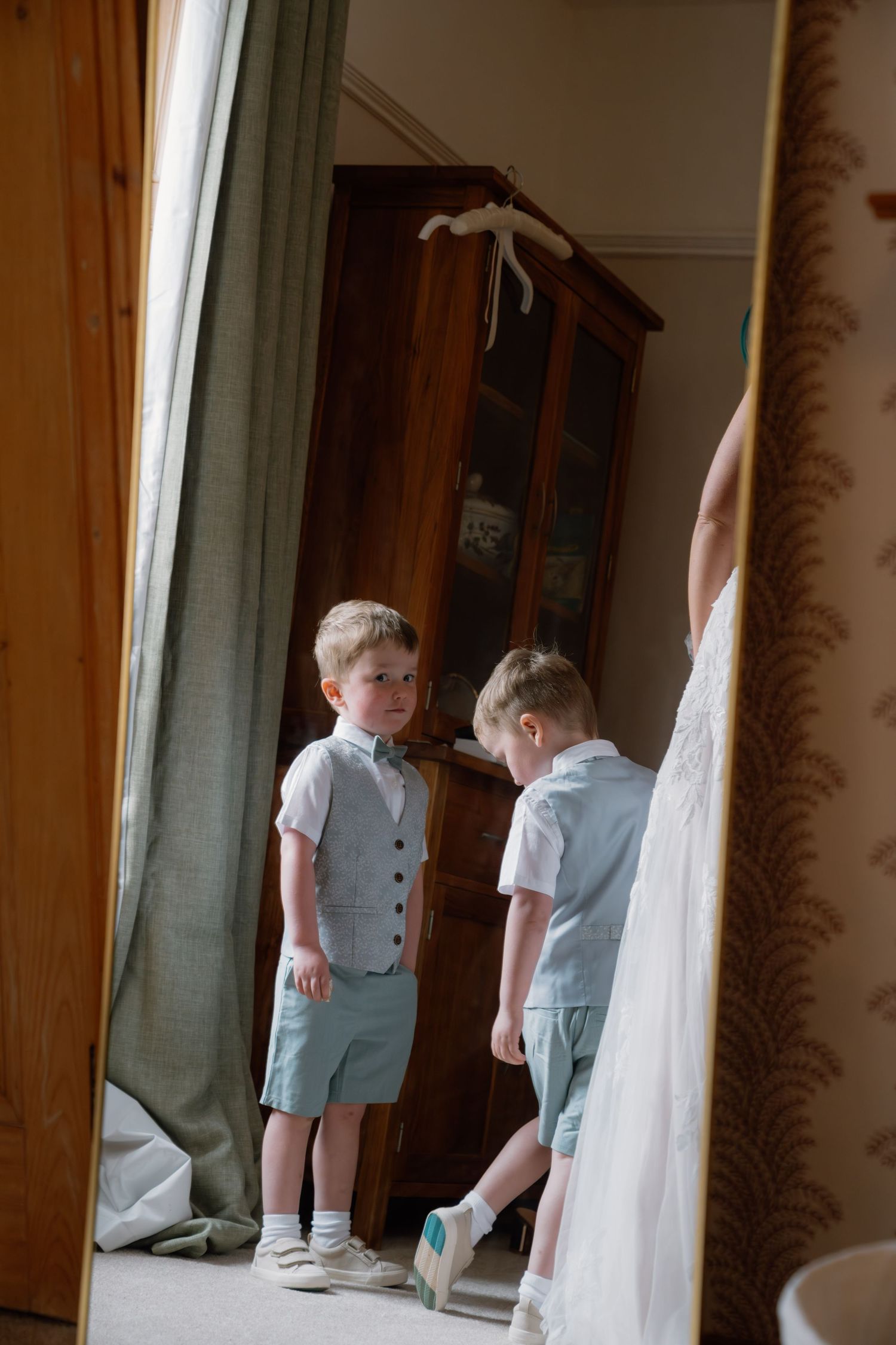 Young ring bearer in gray vest and shorts admires himself in a large mirror during wedding preparations.